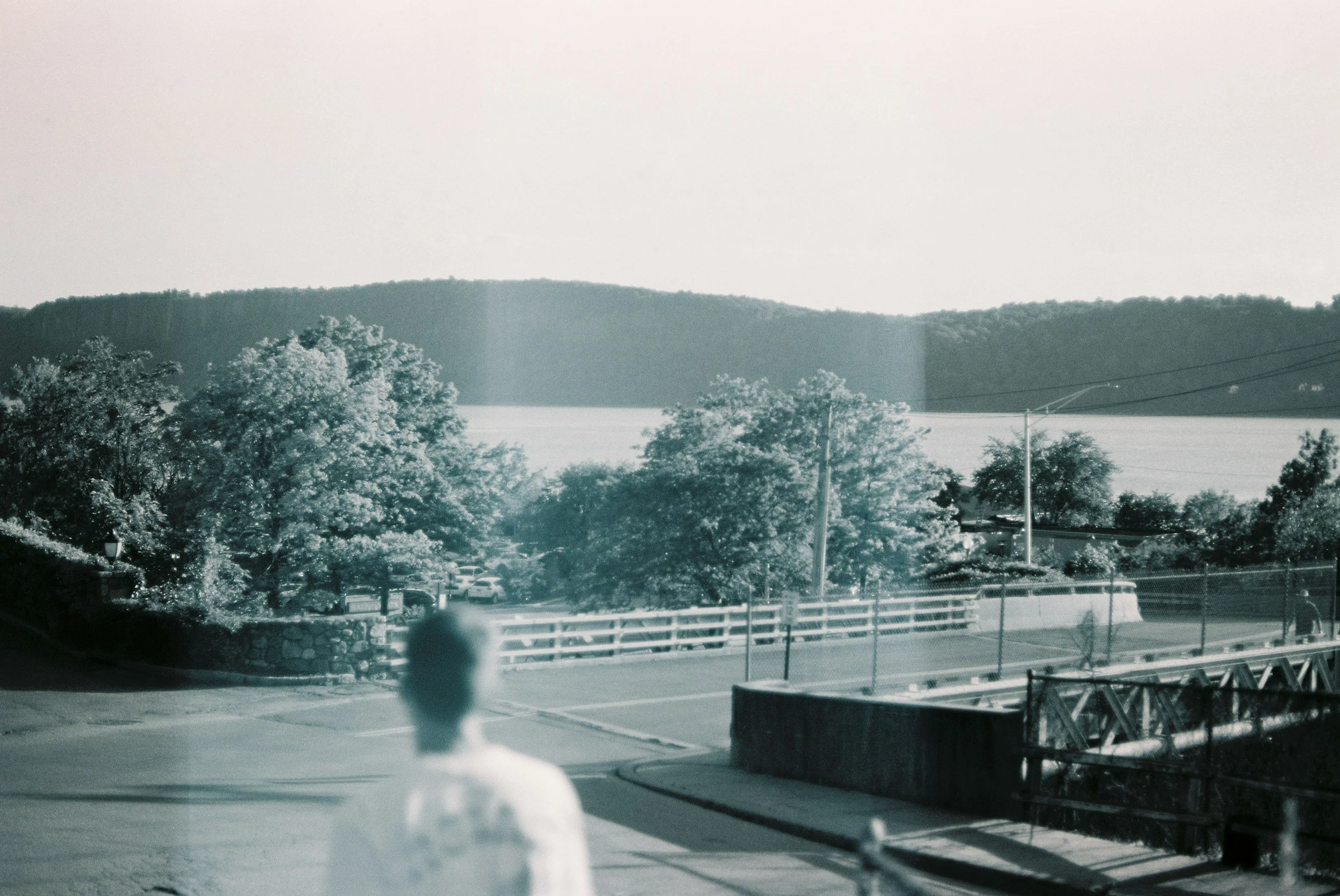 A black-and-white photo of a lakeside scene with hills in the distance, trees in the foreground, and a parking area with cars, as seen through a window with wires and a person in the bottom left corner.