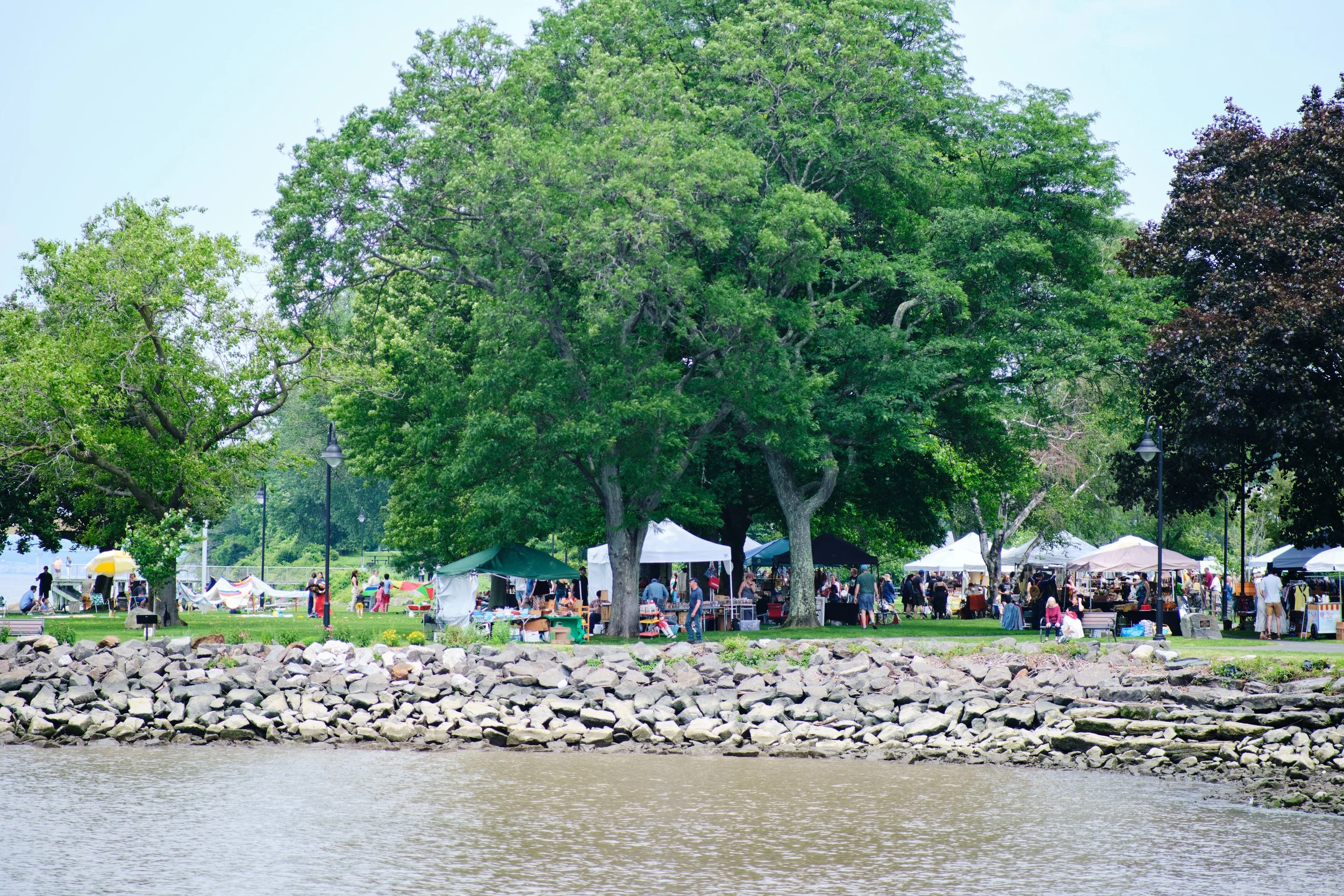 Park with large trees, a grassy area, and outdoor market tents, along a rocky shoreline by a body of water, with people walking and browsing.