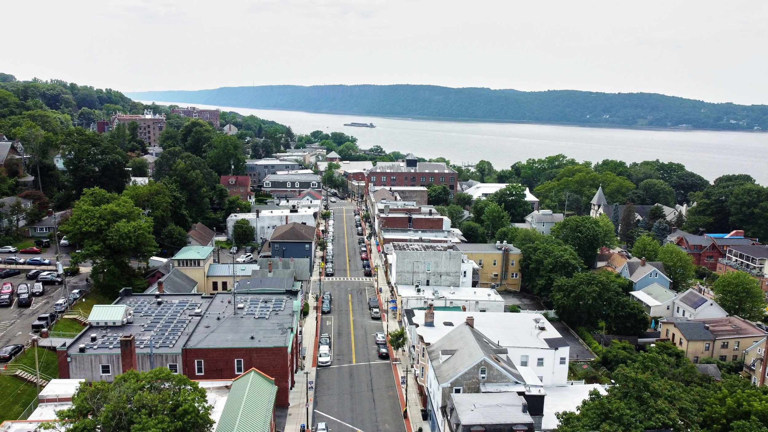Aerial view of a small town by a large river, with buildings, trees, and a boat on the water.