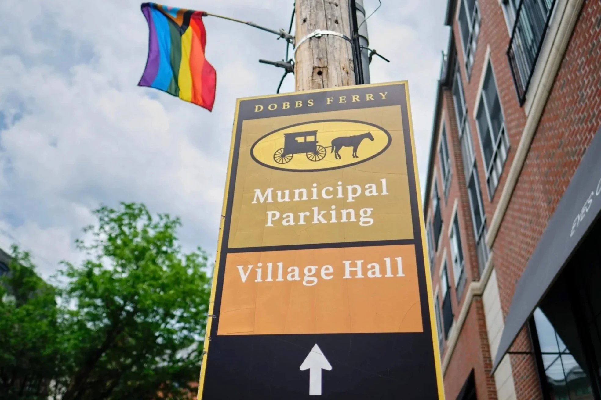 A street sign for Dobbs Ferry with directions to municipal parking and village hall. A rainbow pride flag is attached to a utility pole next to the sign, against a background of a brick building and trees with green leaves, under a partly cloudy sky.