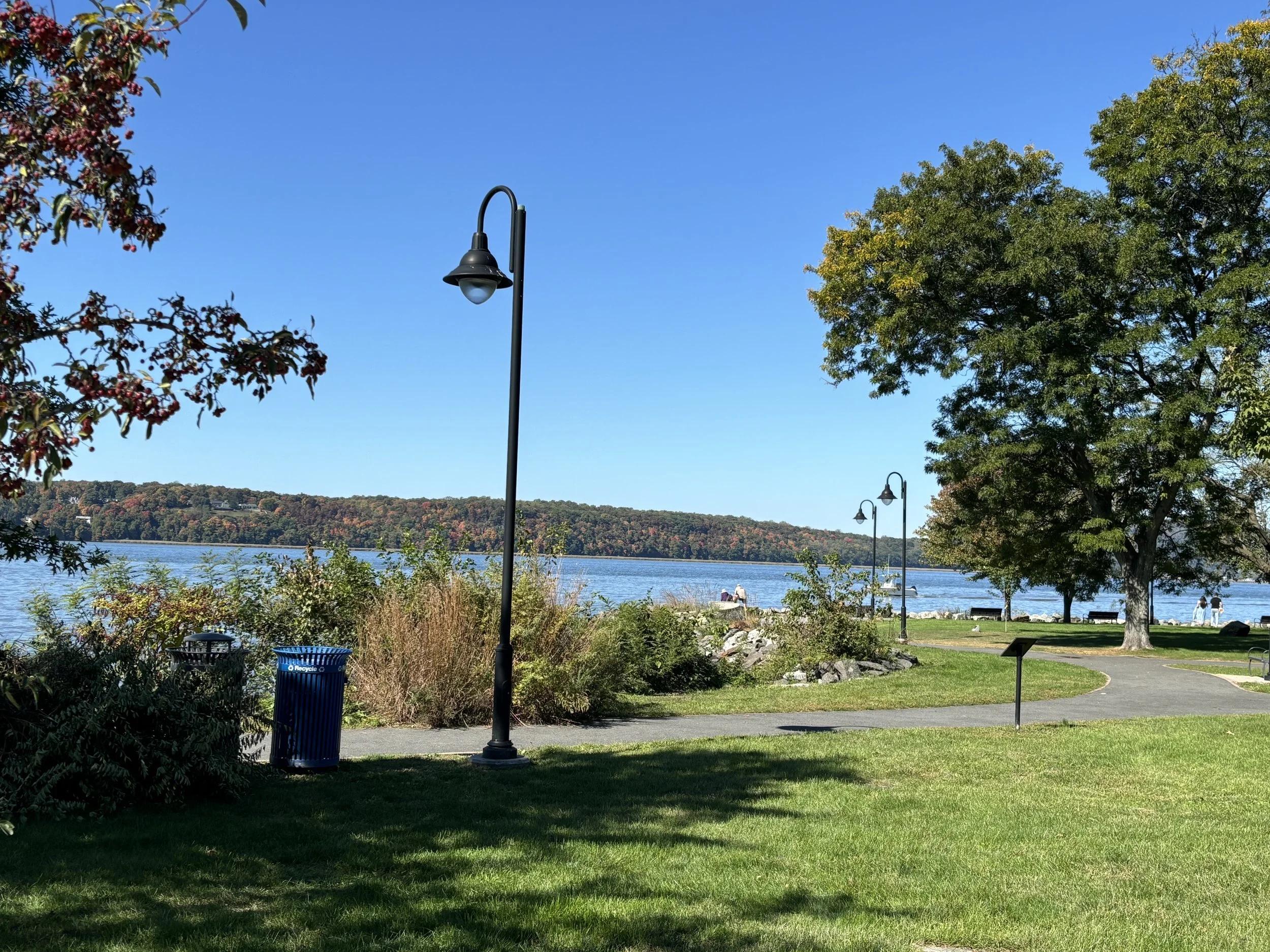 A lakeside park with walking paths, tall trees, benches, lamp posts, a trash bin, and a view of a large body of water with a distant hilly landscape covered in fall foliage under a clear blue sky.