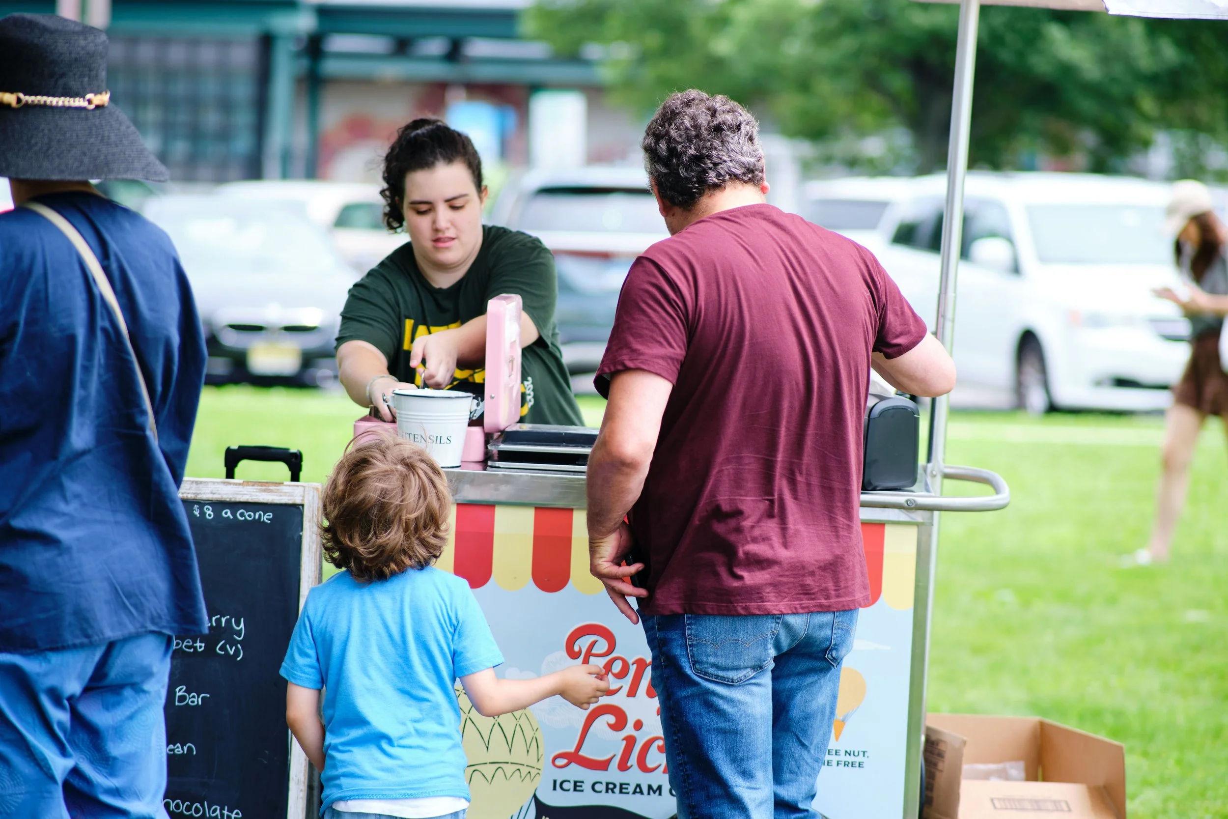 A woman serving ice cream at a festival or fair, with two children and a man in line. The woman is behind a colorful ice cream cart, and there is a blackboard with prices and flavors listed.