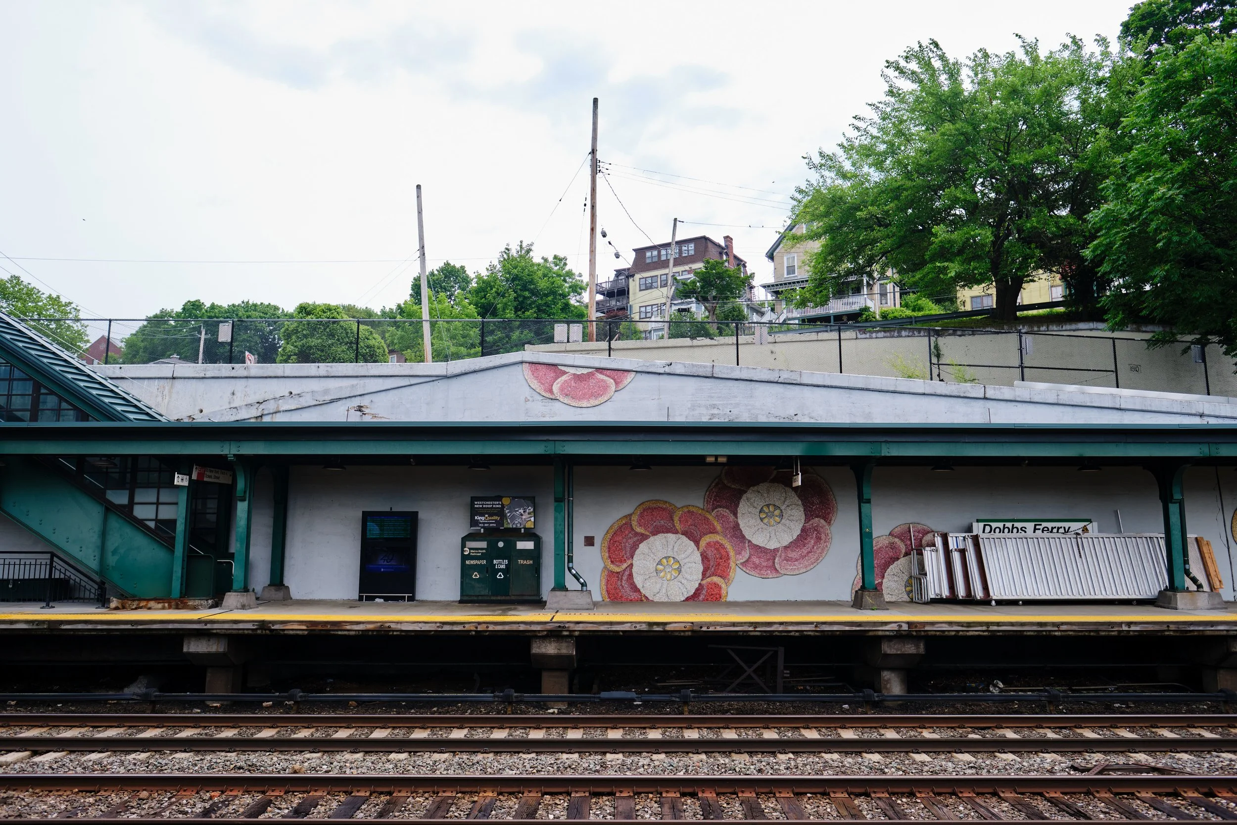 View of a train station platform, with railway tracks in the foreground and a wall with floral mural and digital information boards above. In the background, a second level with residential houses, trees, and utility poles.