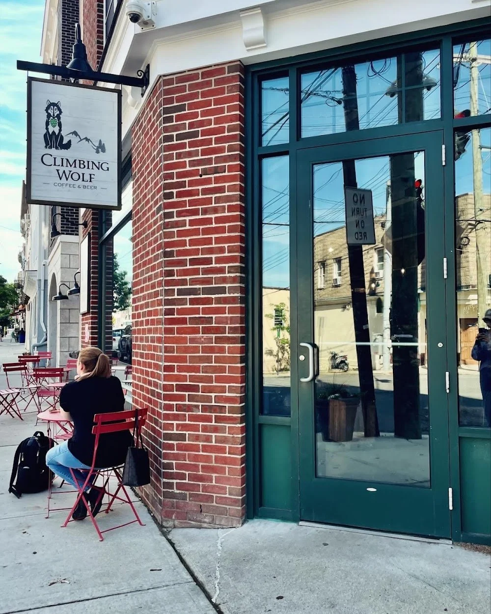 Exterior of Climbing Wolf coffee and beer shop with a woman sitting outside at a red table on a sidewalk, red chairs, and a brick building with large glass door and window reflections.