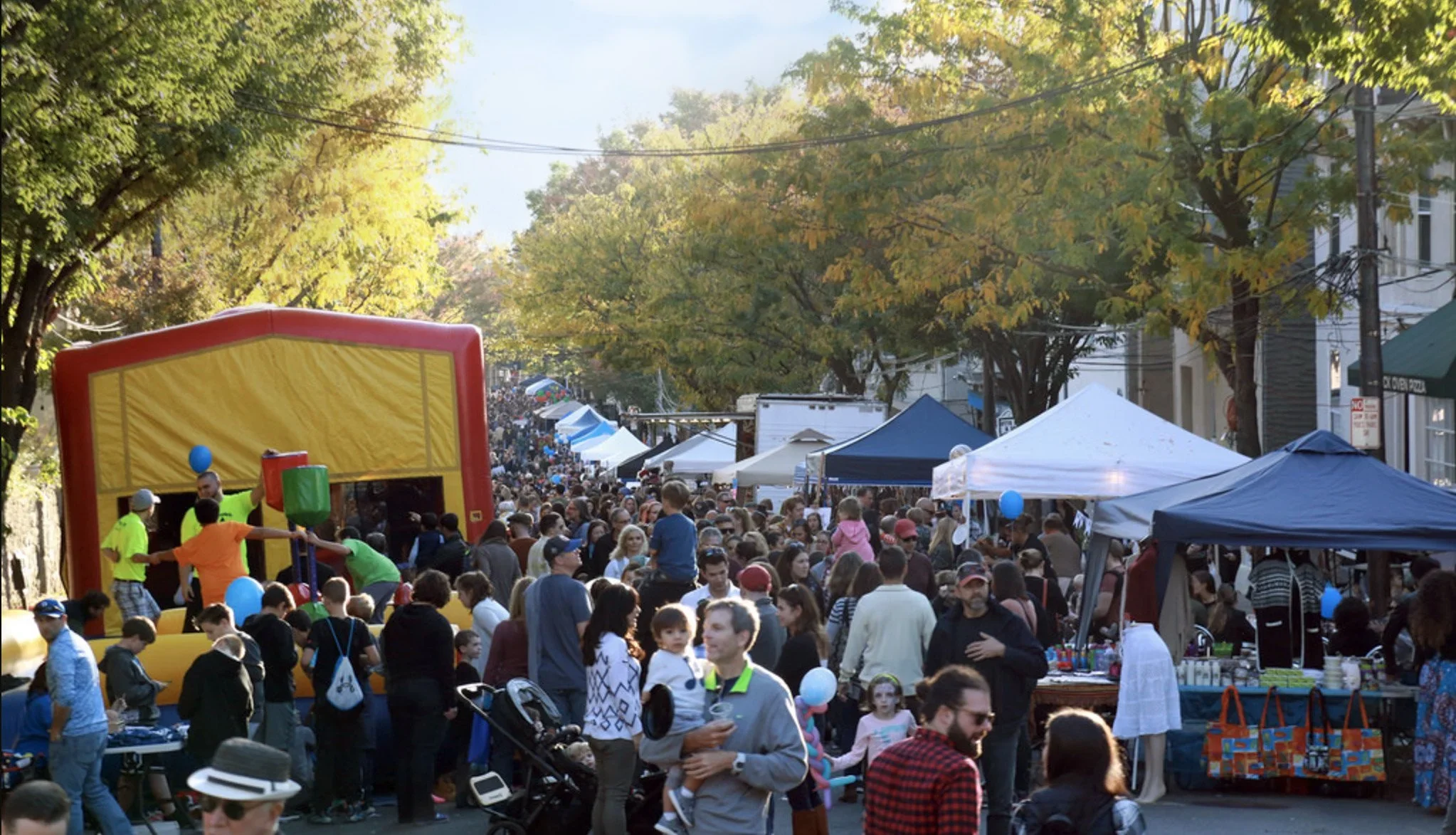 A street fair with numerous tents and booths lining the street, filled with a large crowd of people. The scene is shaded by trees with green and yellow leaves, indicating autumn. There are children on a yellow inflatable bounce house, and various vendors and attendees enjoying the event.