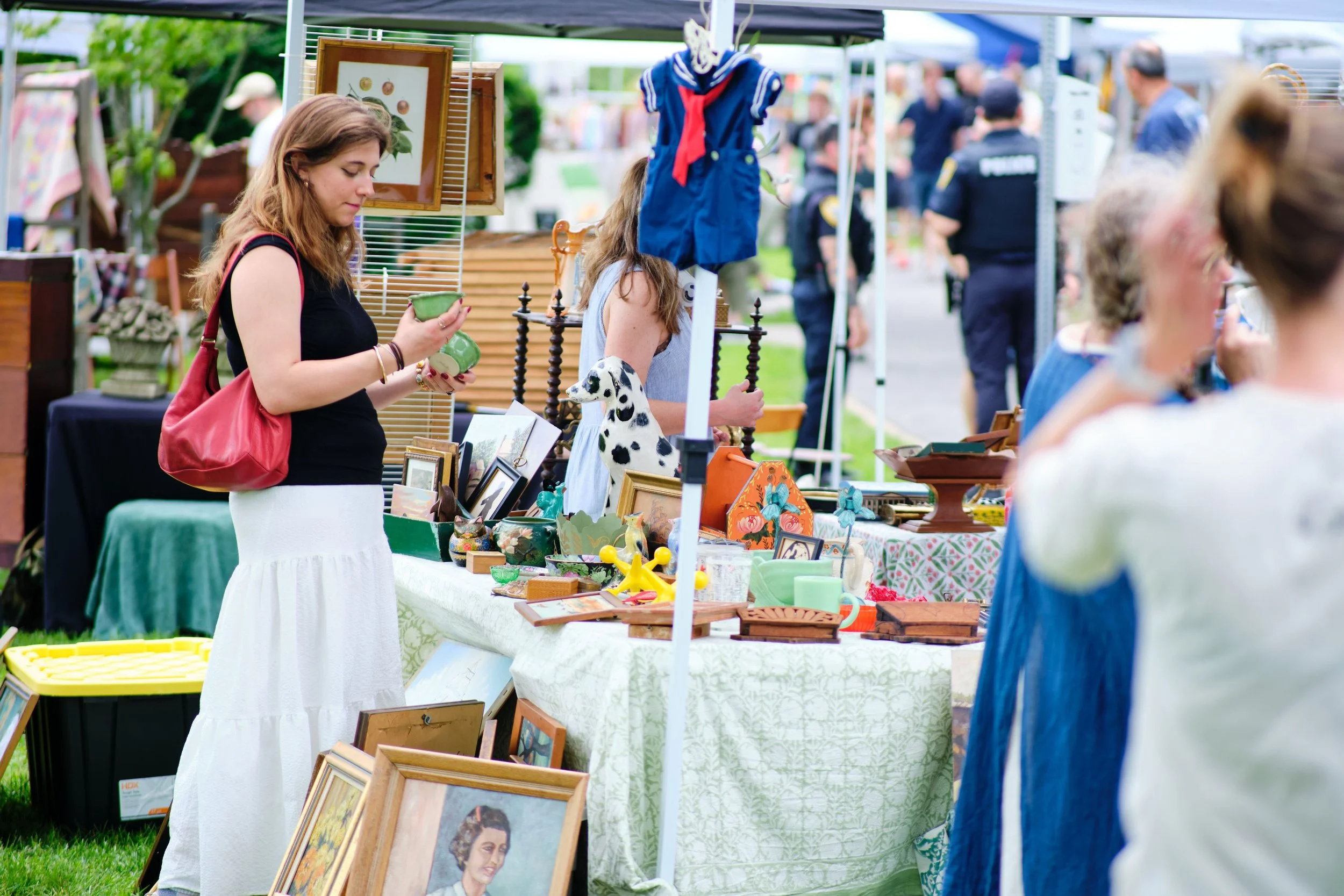 People browsing at an outdoor art and craft fair with tables displaying paintings, ceramics, and framed art.
