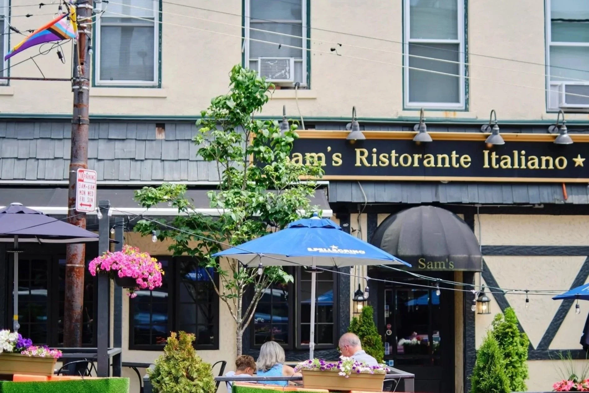 Outdoor dining area in front of Sam's Ristorante Italiano, with umbrellas, potted flowers, and people sitting at tables.