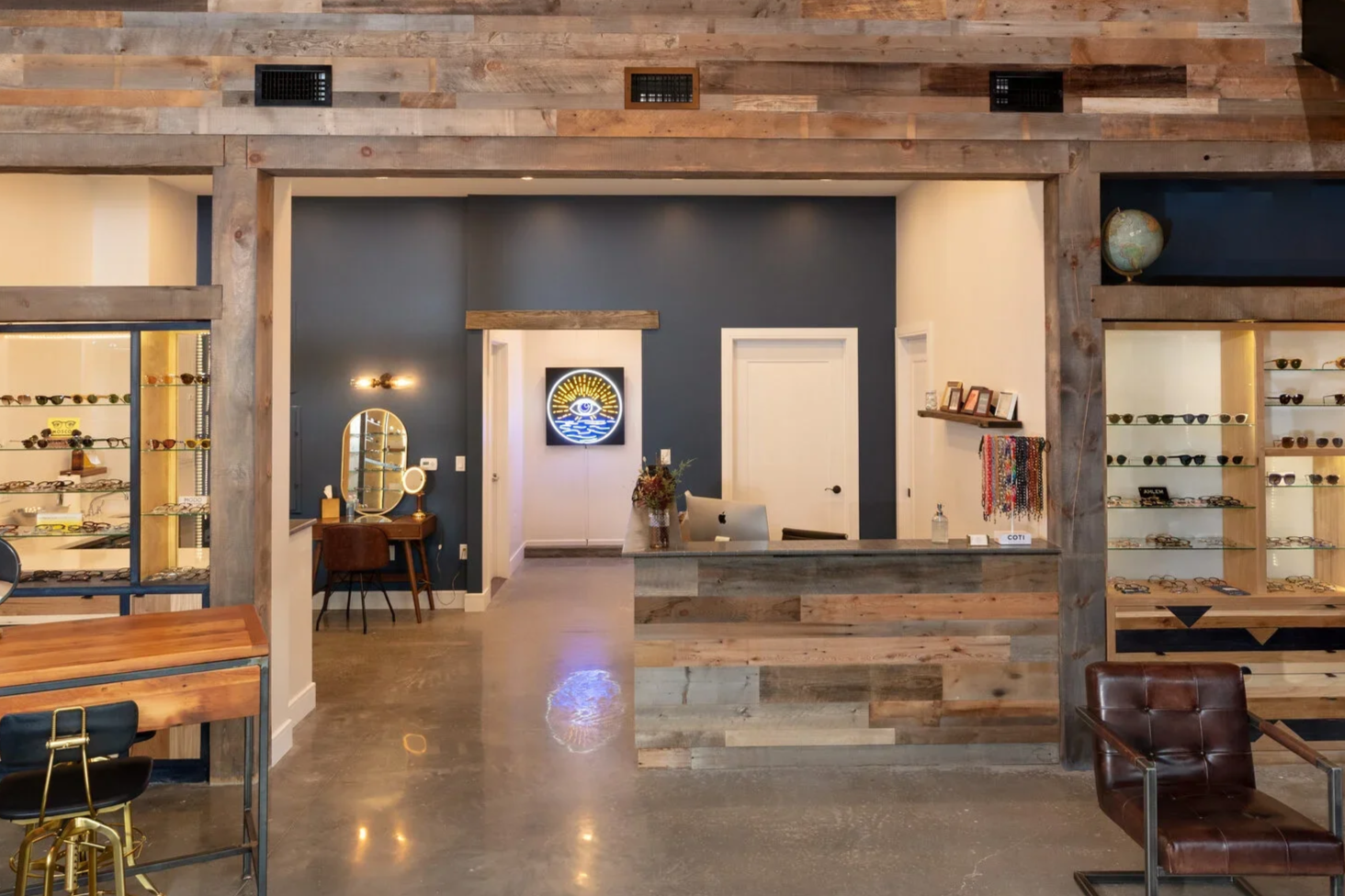 Interior of a boutique eyewear shop with display shelves of sunglasses, a reception counter, and a seating area, featuring a mix of modern and rustic decor with wood accents and a polished concrete floor.