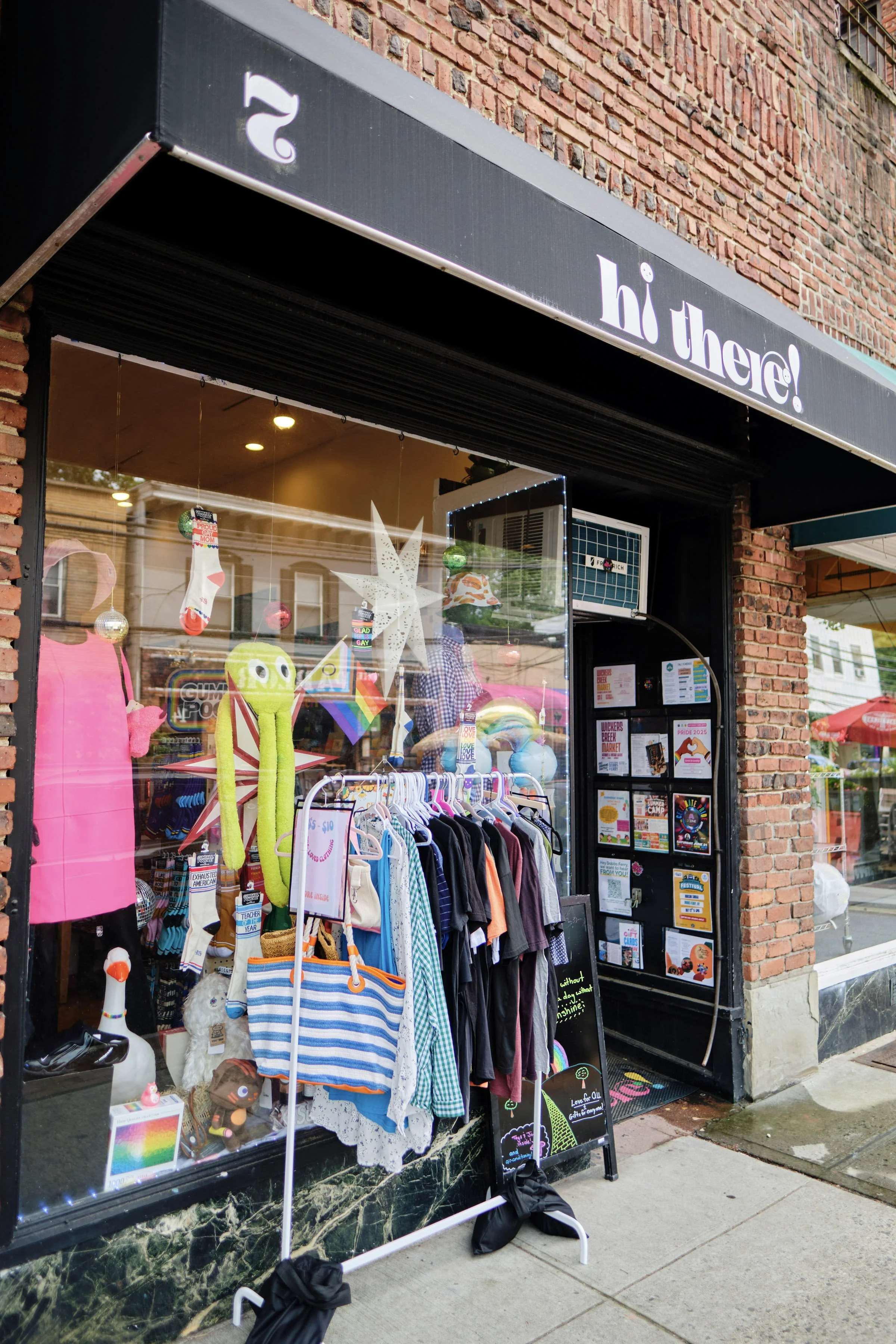 A storefront display with clothing items, plush toys, and rainbow-themed accessories inside a window. A variety of signs and flyers are posted near the entrance, and the store has a black awning with the words 'hi there!' in white. The exterior features red brick walls.