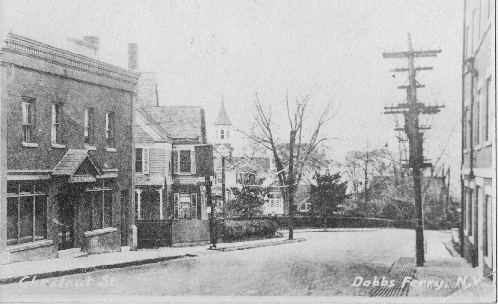 Old black and white photo of a street corner with brick buildings, leafless trees, and a utility pole in the foreground. There is a church steeple in the background and the street names 'Main Street St' and 'Dobbs Ferry' are visible.