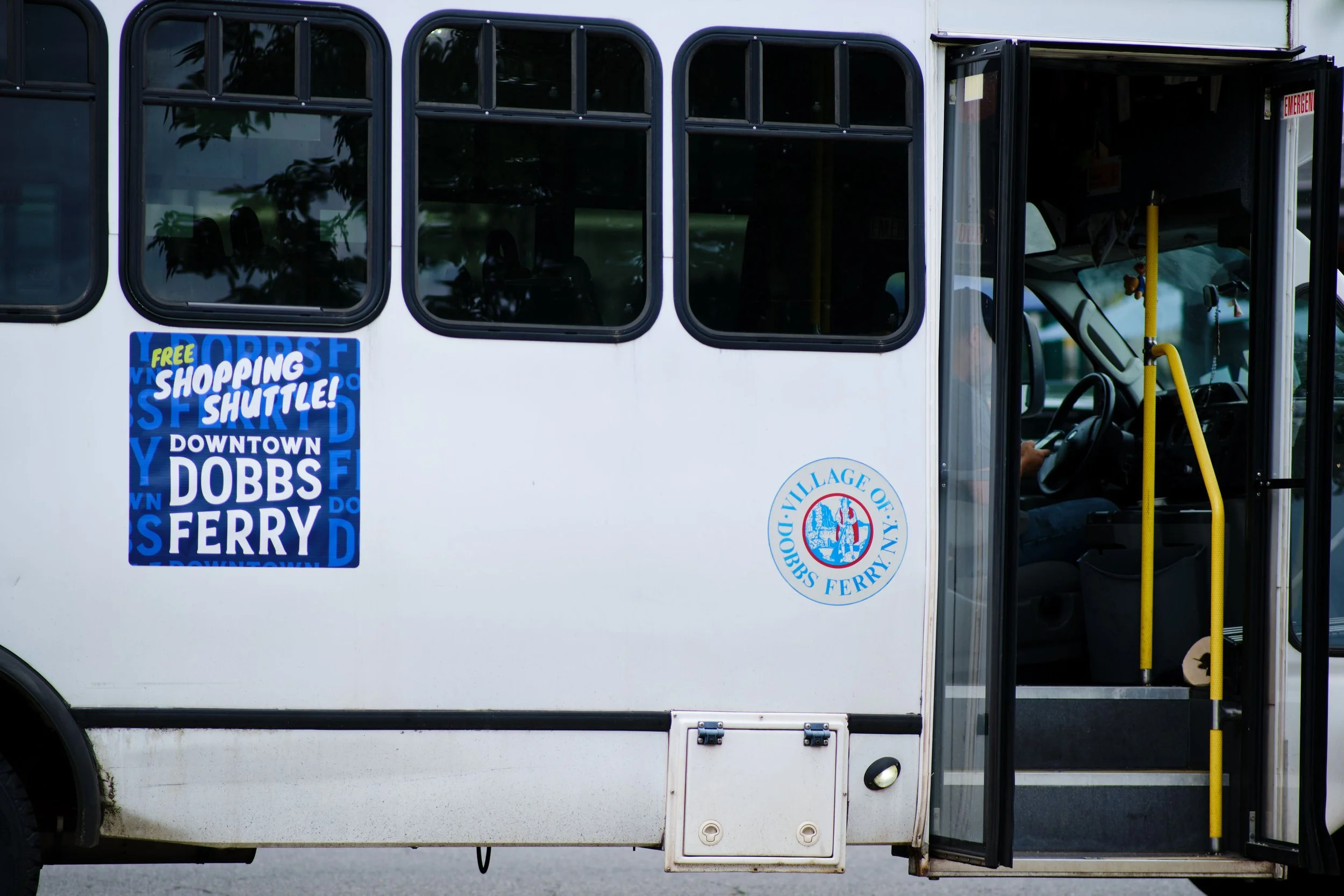 A white shuttle bus with black windows and a yellow pole near the open door, showing the driver inside. There is a blue sign advertising free shopping shuttle service to Downtown Dobbs Ferry, and a circular emblem of the Village of Dobbs Ferry on the bus.