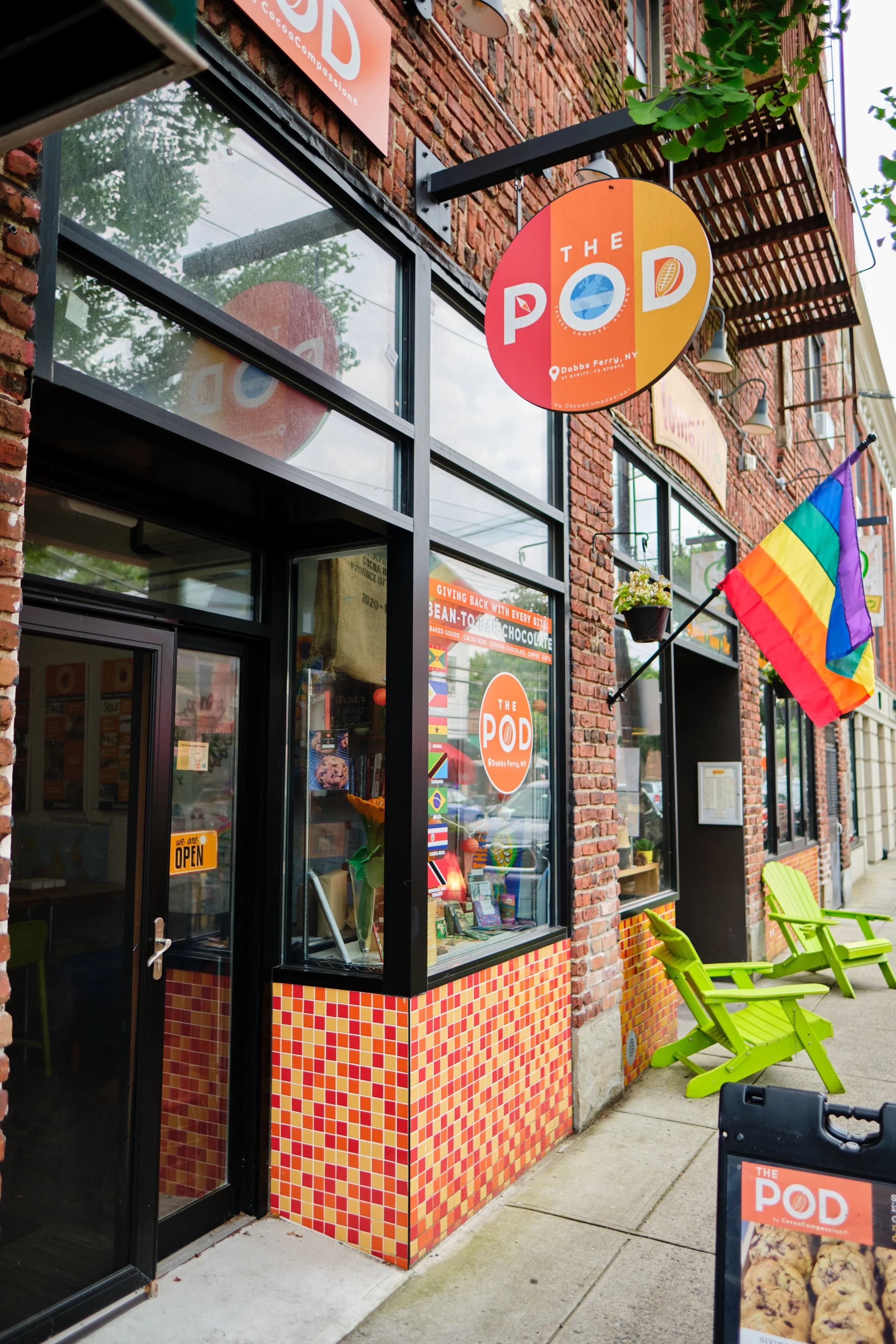 The storefront of a shop called 'The Pod' located at Dobbs Ferry, NY, with a rainbow pride flag outside, green chairs on the sidewalk, and a large window displaying posters and signs.