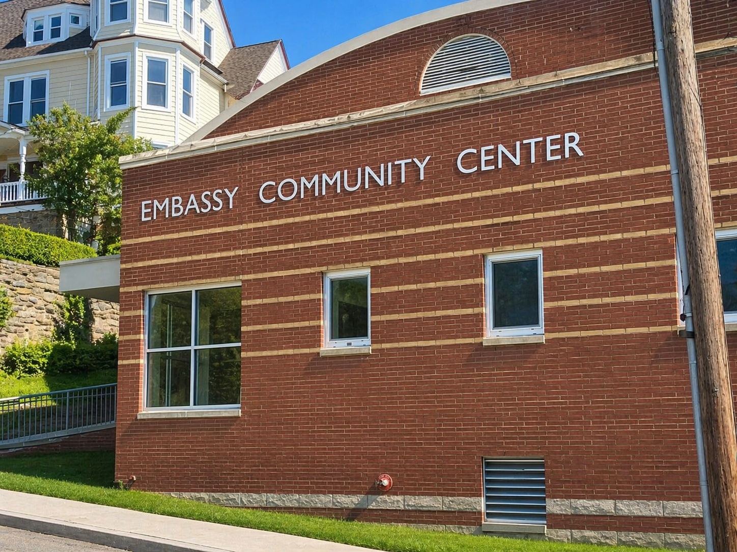 Red brick building with white sign reading 'Embassy Community Center' on the front, multiple windows, a grassy area, a sidewalk, and residential houses in the background under a blue sky.