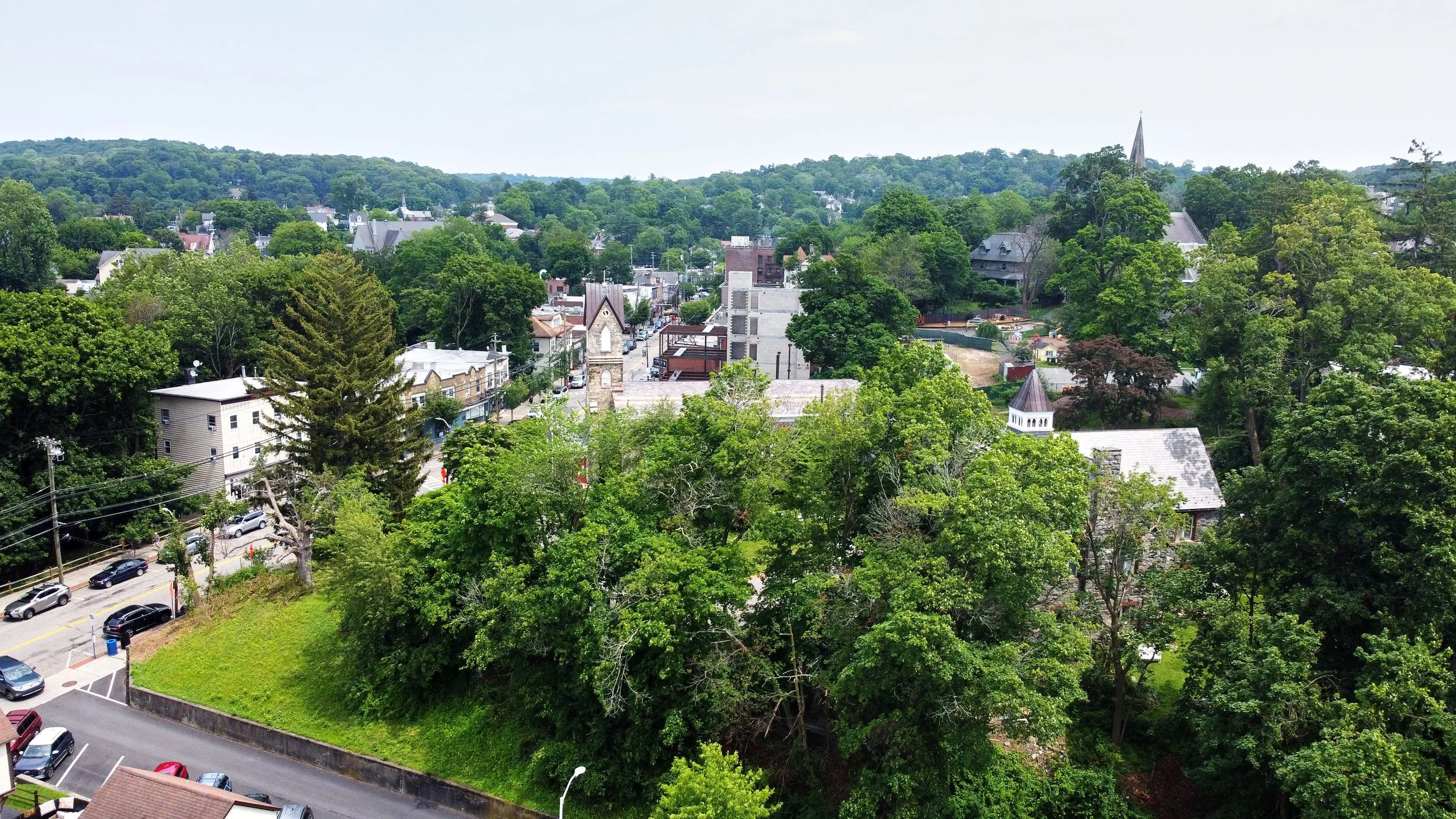 Aerial view of a small town with many trees, buildings, and streets, with a hill in the background and a steeple of a church visible.