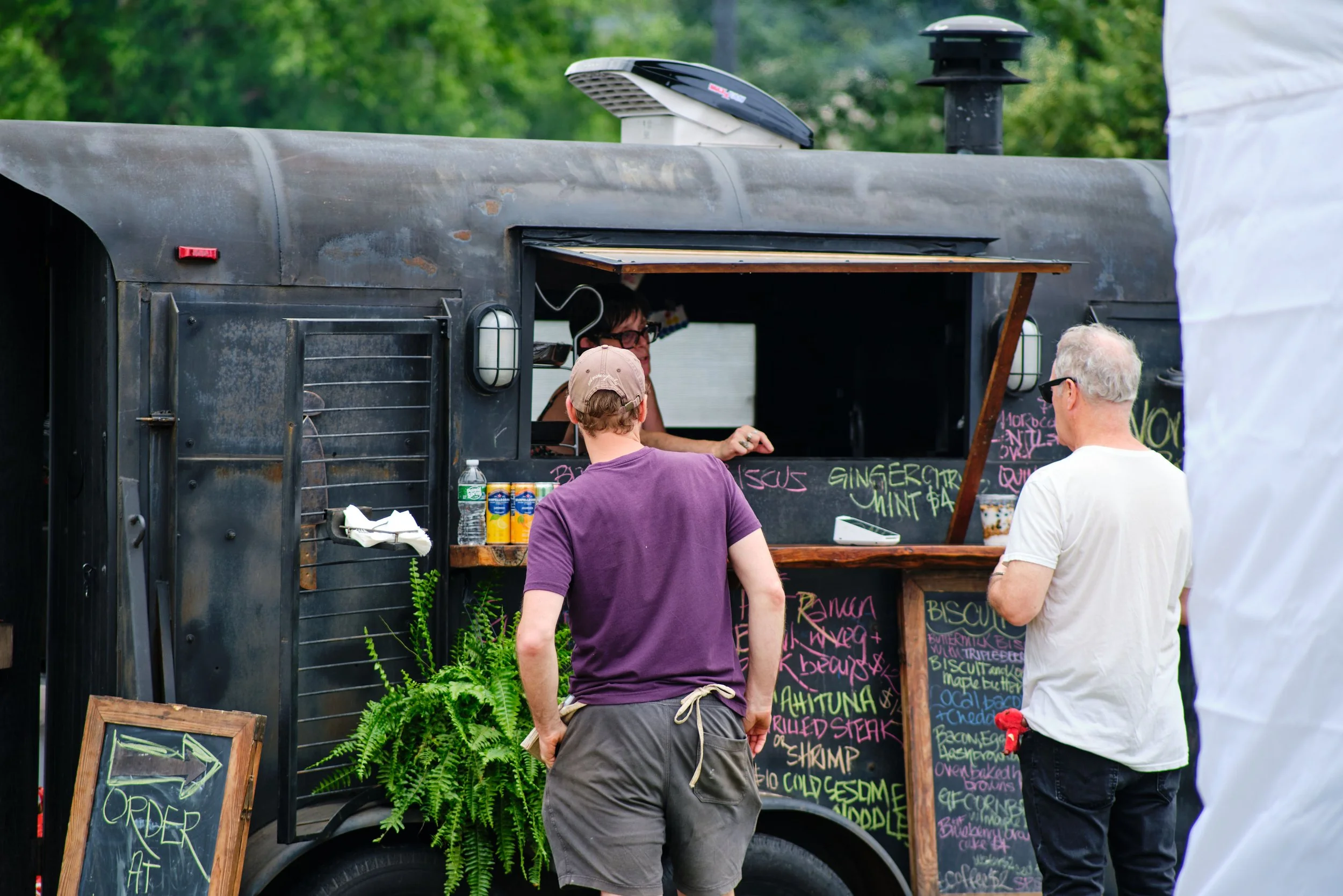 People ordering food at a black food truck with colorful menu chalkboards in an outdoor setting with green trees in the background.