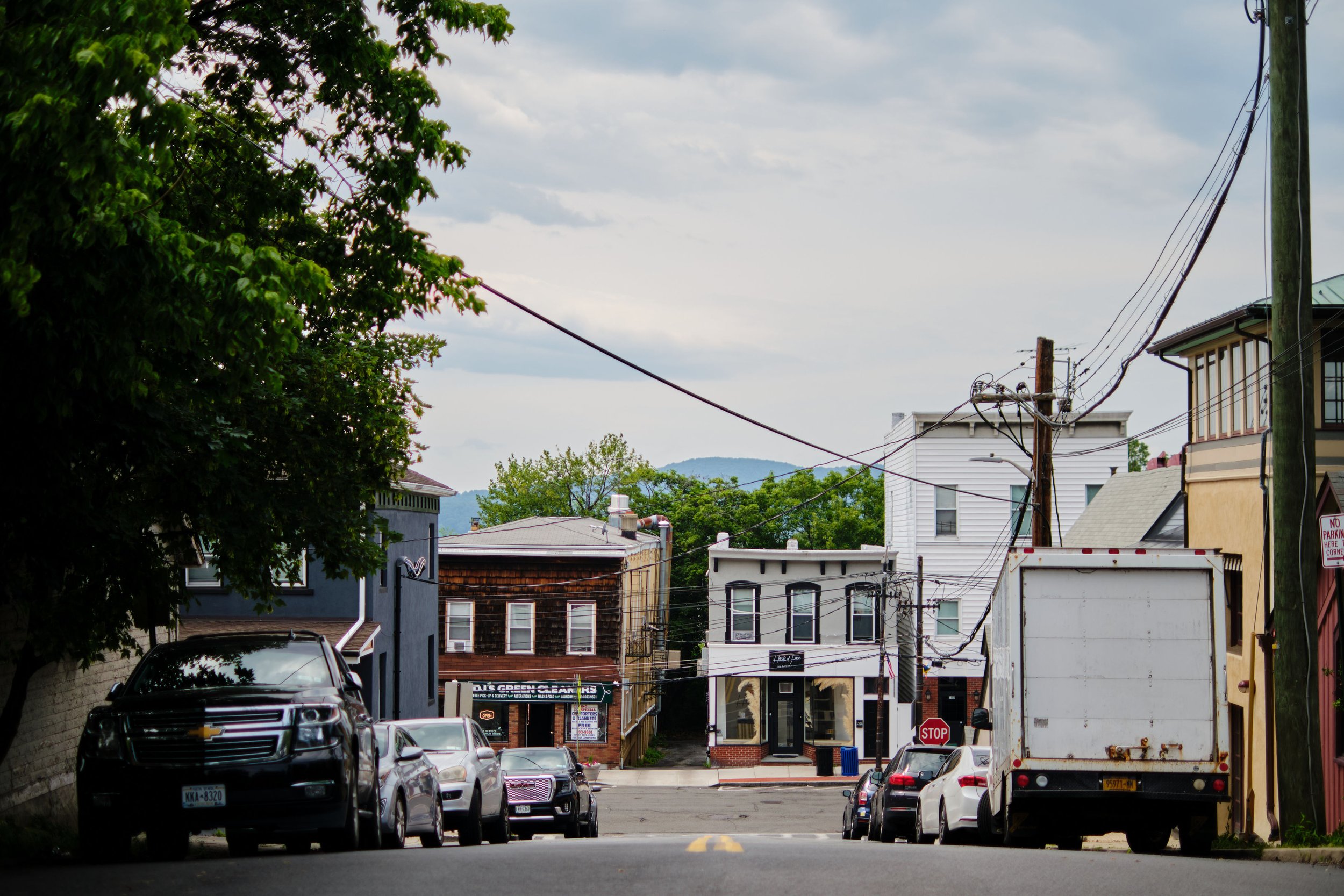 A small town alley with parked cars on both sides, utility poles with wires, and buildings with shops and residential apartments. Trees line the street, and a cloudy sky is above.