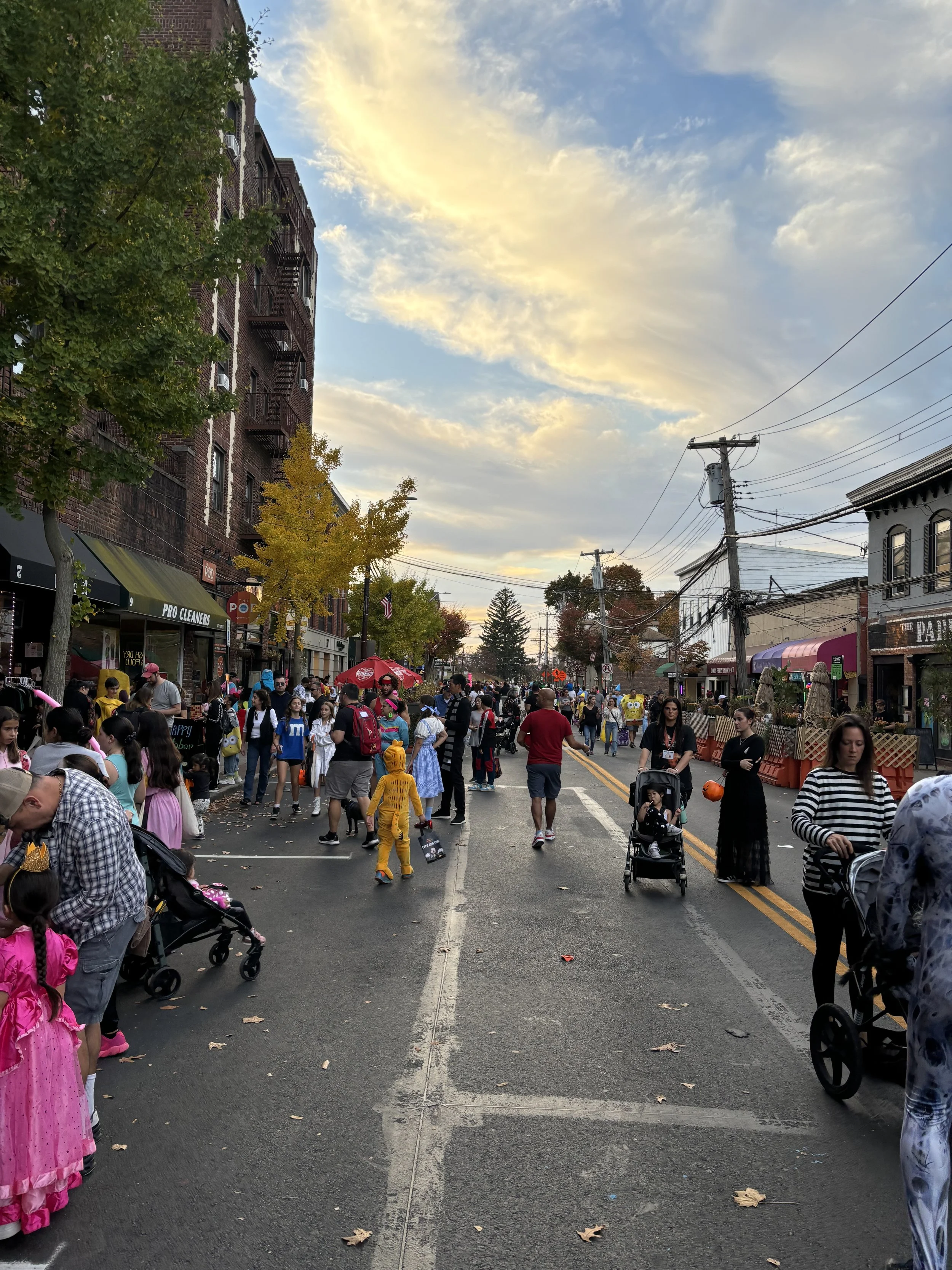 Street scene during daytime with many people walking and socializing, some dressed in costumes, storefronts on each side, trees with yellow and green leaves, cloudy sky with sunlight, utility poles and power lines overhead.