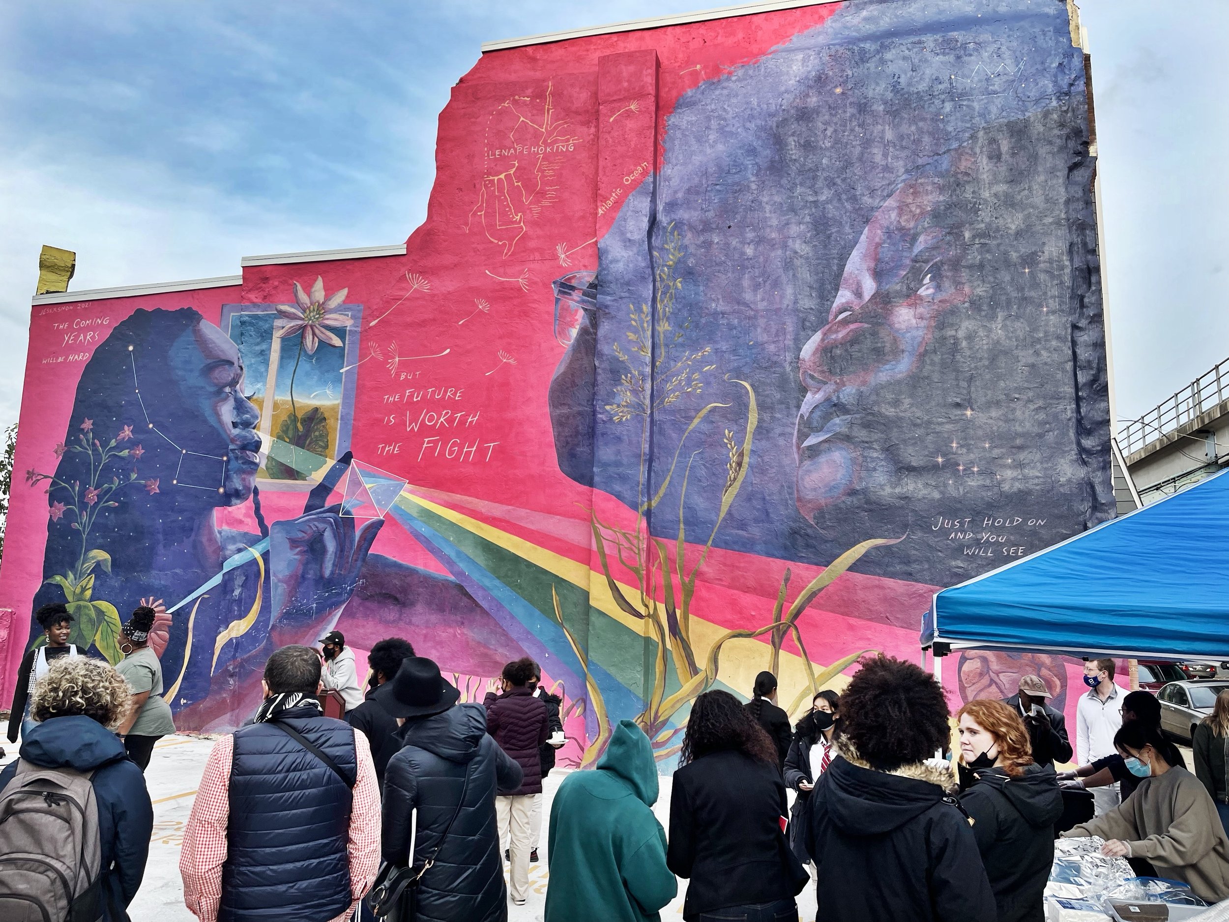 An image of people gathered in front of the unveiling of the JesxSnow mural across the street from J-Centrel. The mural is a vibrant depiction of black femme characters against a bright fuscia colored background and a prism refracting a rainbow.