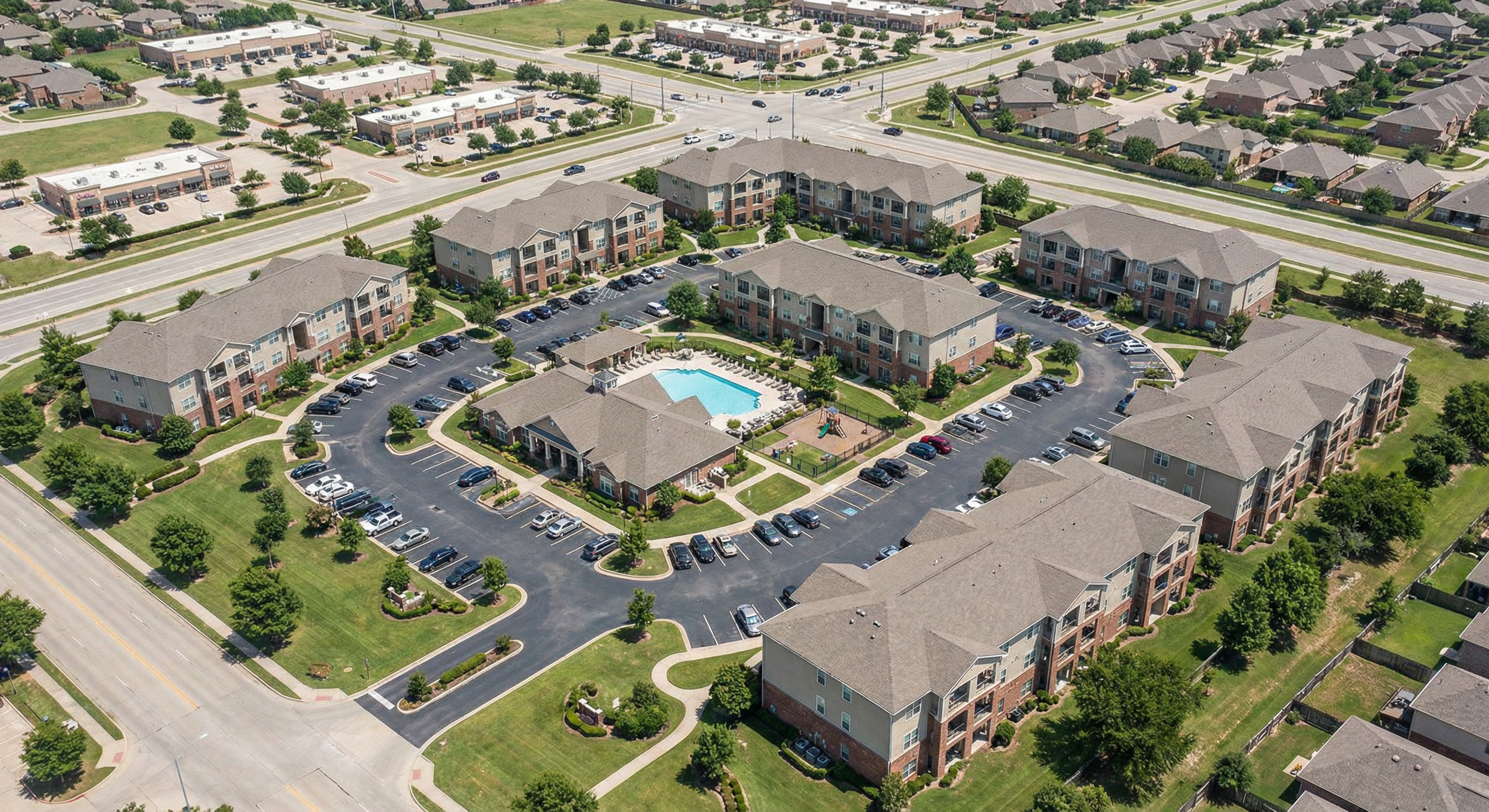 Aerial view of a residential apartment complex with multiple buildings, parking lots, a swimming pool, a playground, and surrounding greenery.