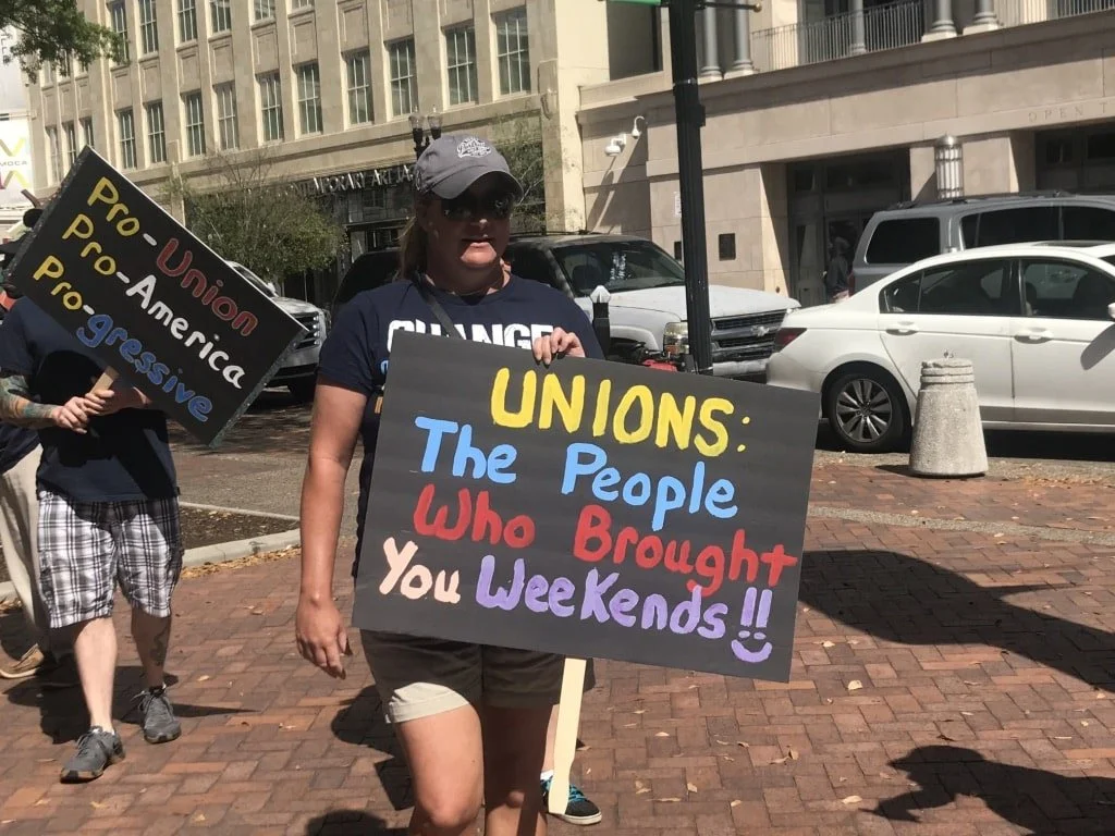 A woman at a protest holding a sign that reads 'Unions: The People Who Brought You Weekends!!' with other protesters holding signs in the background.