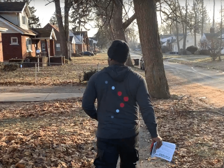 A person wearing a dark hoodie and black pants walking on a leaf-covered sidewalk in a residential neighborhood, holding a clipboard, with houses and trees in the background under a sunny sky.