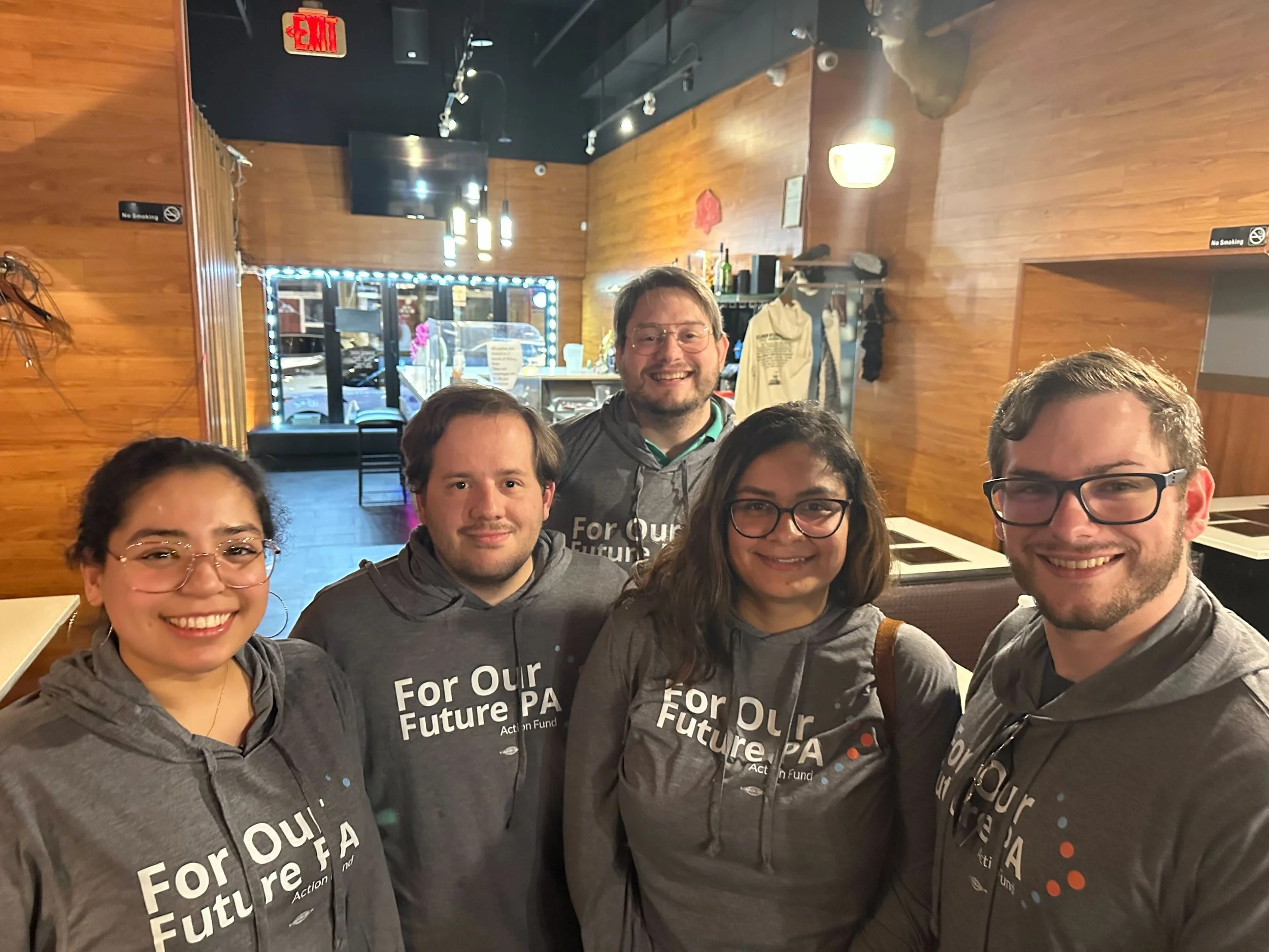 Group of five young adults smiling inside a restaurant or cafe, wearing matching gray hoodies with 'For Our Future PA Action Fund' printed on them.