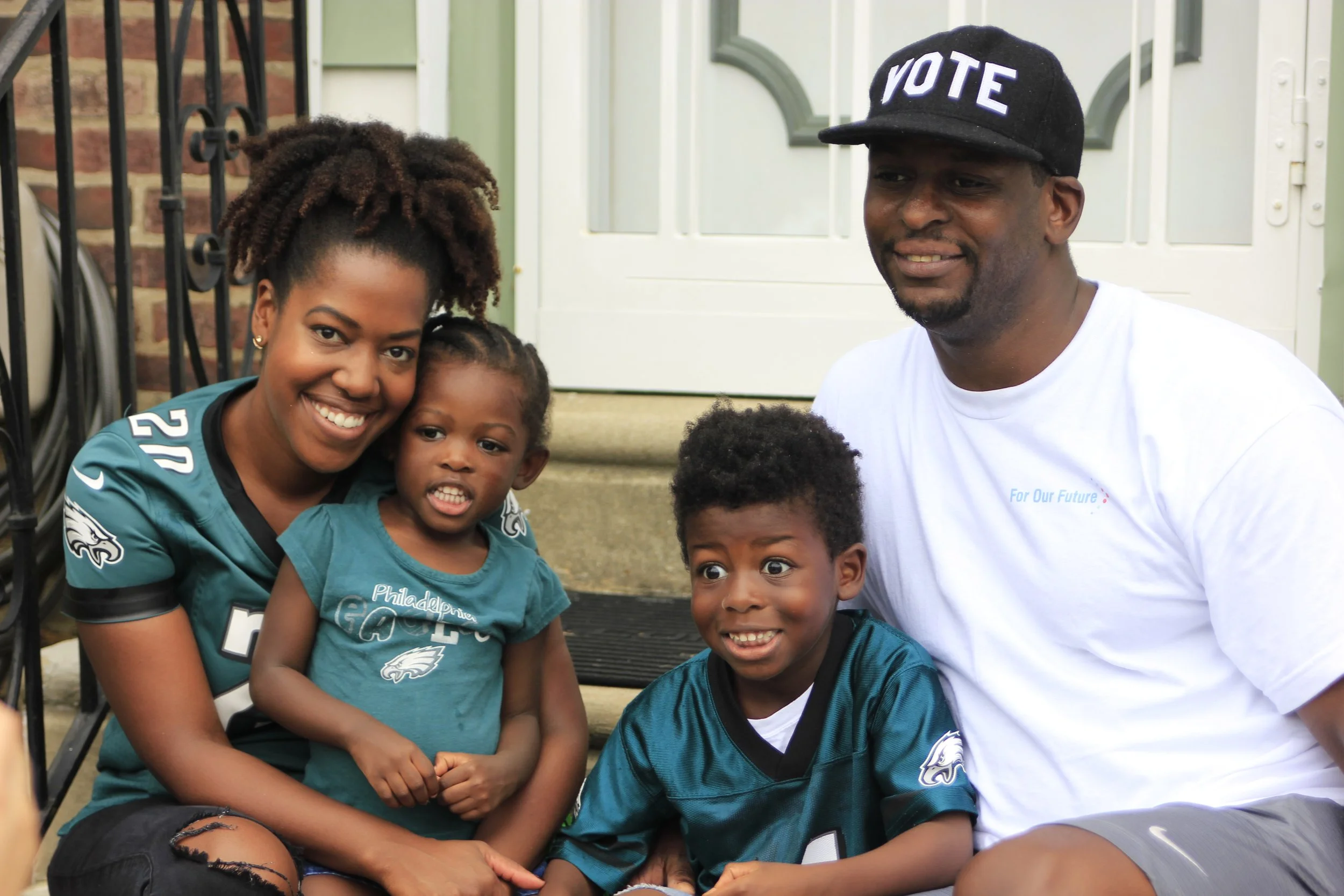 A smiling woman and man with children outdoors on a porch, all wearing Philadelphia Eagles football jerseys and shirts, in front of a white door and railing.