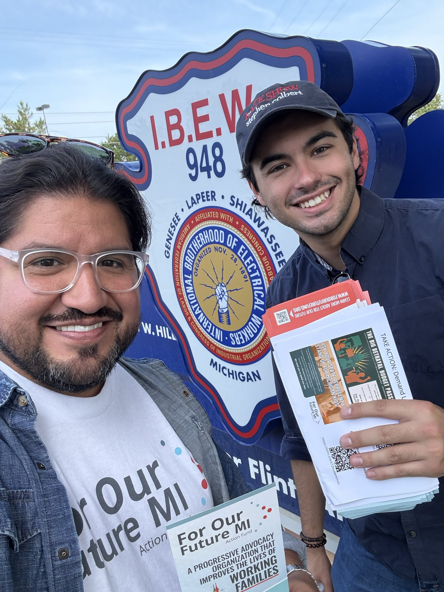 Two people smiling at a rally, holding flyers and standing in front of a sign for the International Brotherhood of Electrical Workers in Michigan.