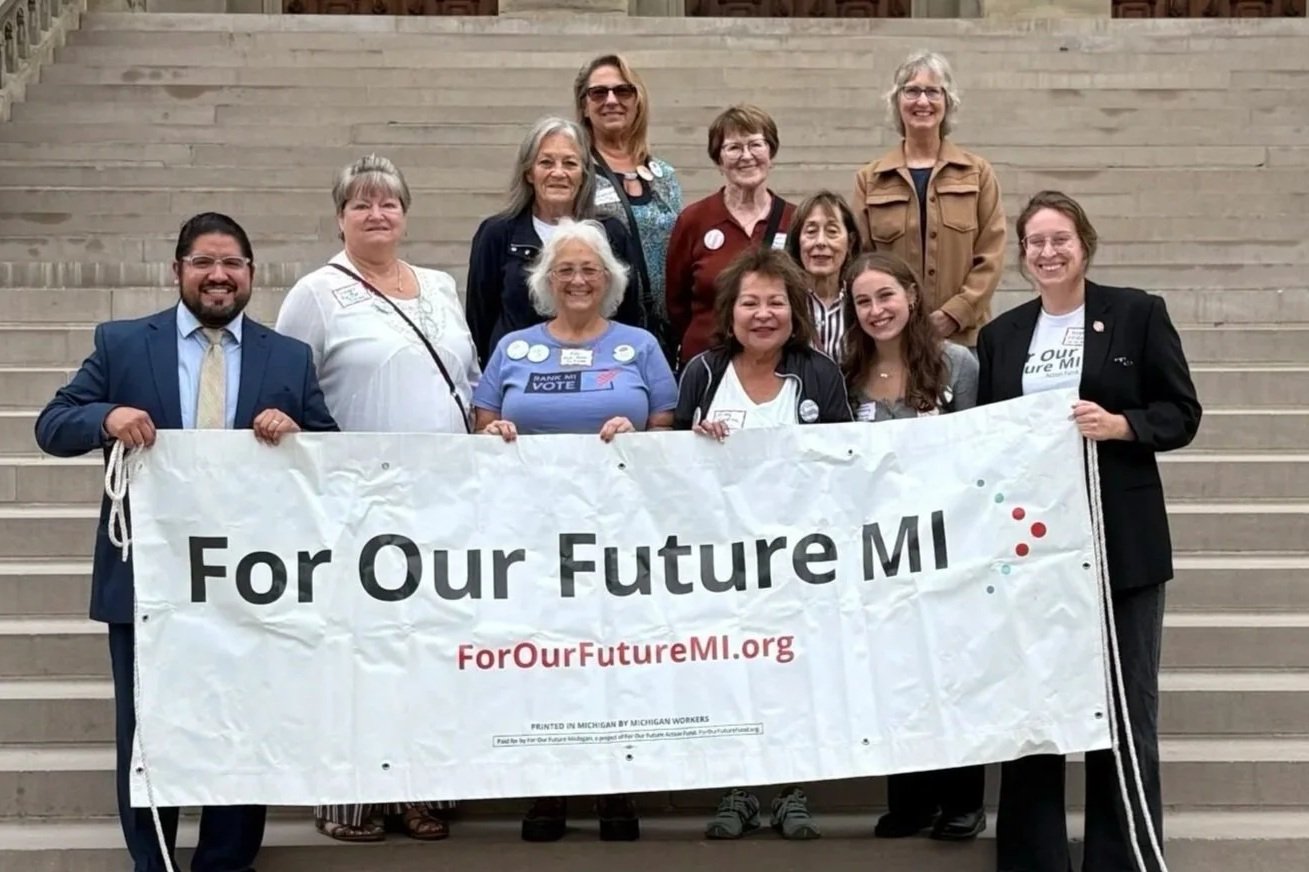 A group of thirteen diverse people on stairs holding a banner that reads 'For Our Future MI' with the website 'ForOurFutureMI.org', smiling and participating in an advocacy or campaign event.