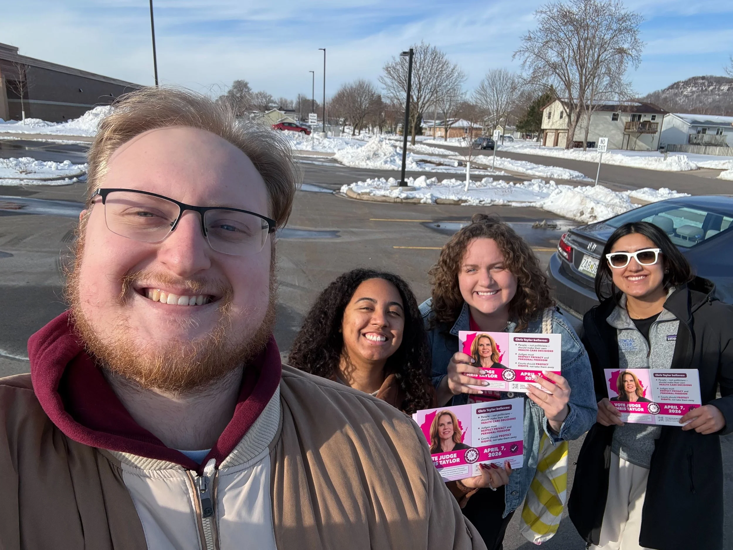 A group of people standing outdoors in a parking lot during winter, holding flyers and smiling. Snow covers the ground and trees, with houses and a street in the background.