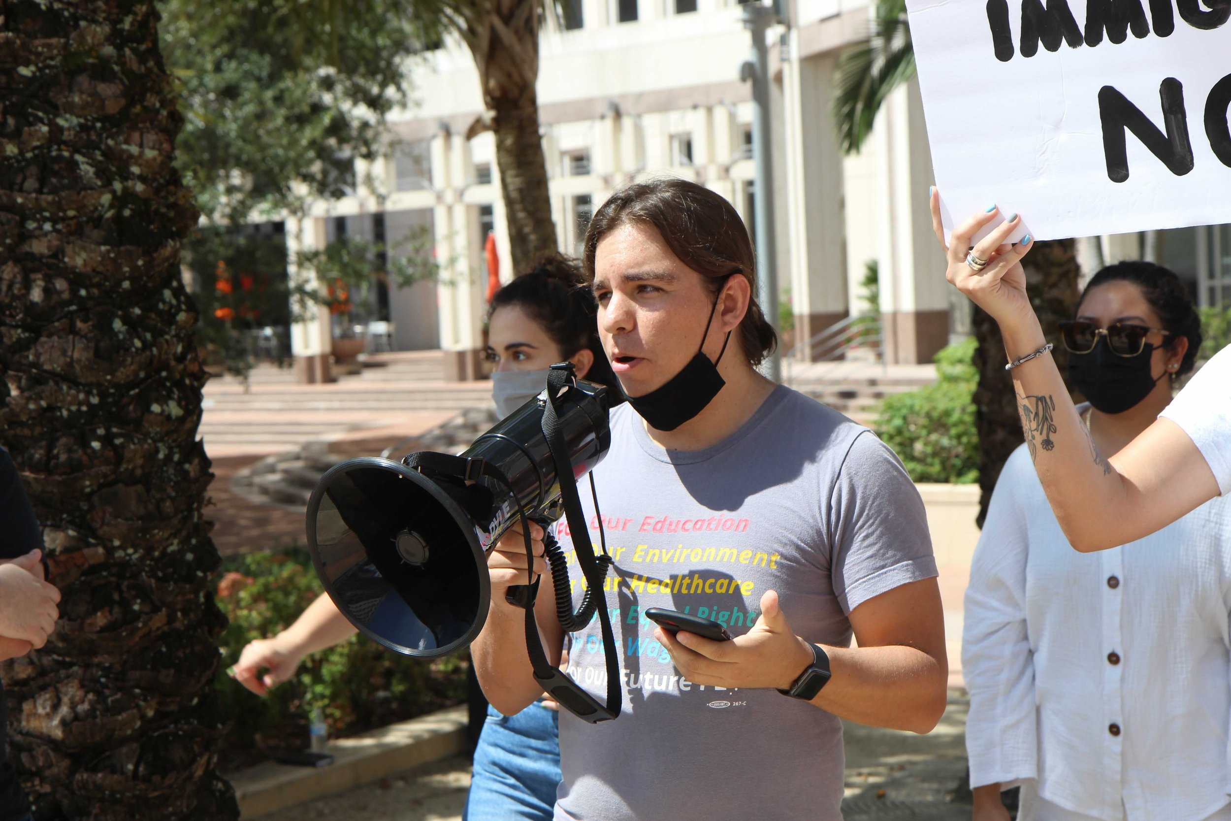 A person with brown hair speaking into a megaphone during a protest or rally, wearing a gray t-shirt with text and a black face mask pulled down below their chin, while holding a smartphone in their hand. Two other individuals, one with glasses and tattoos and another with a mask, are visible in the background, along with trees and a building.