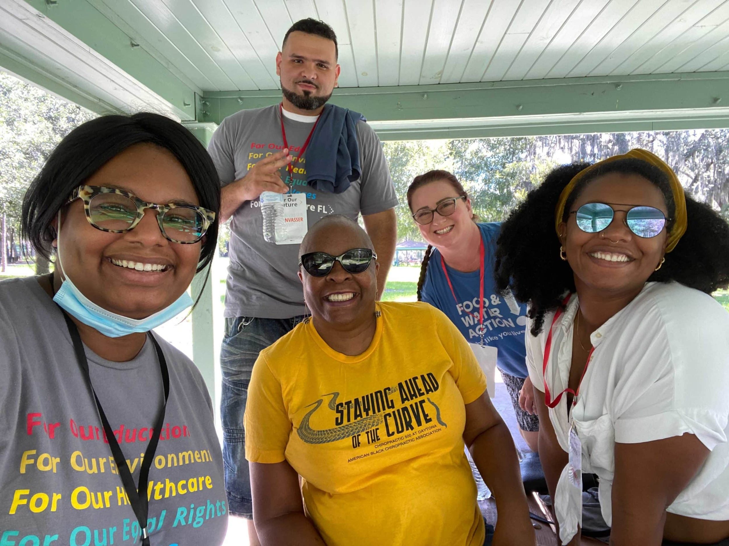 Group of six diverse smiling people posing outdoors under a pavilion, some wearing bright T-shirts with advocacy messages, one person with a mask pulled down, in a sunny park setting.