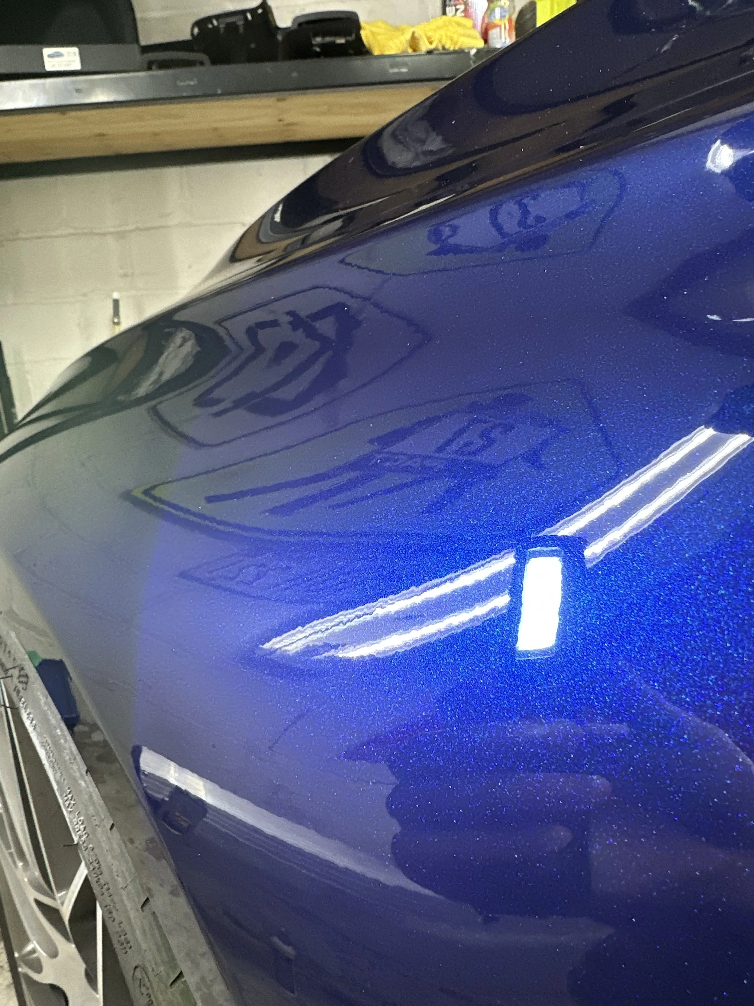 Close-up of a shiny, metallic blue car's side panel with visible reflections and a partially visible tire and wheel.