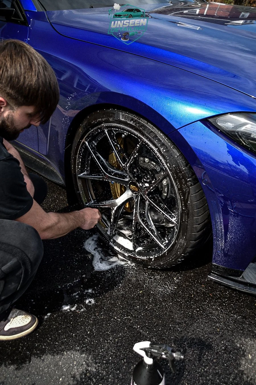 A person cleaning a blue sports car's black alloy wheel with soap, water, and a sponge during a car detailing session.