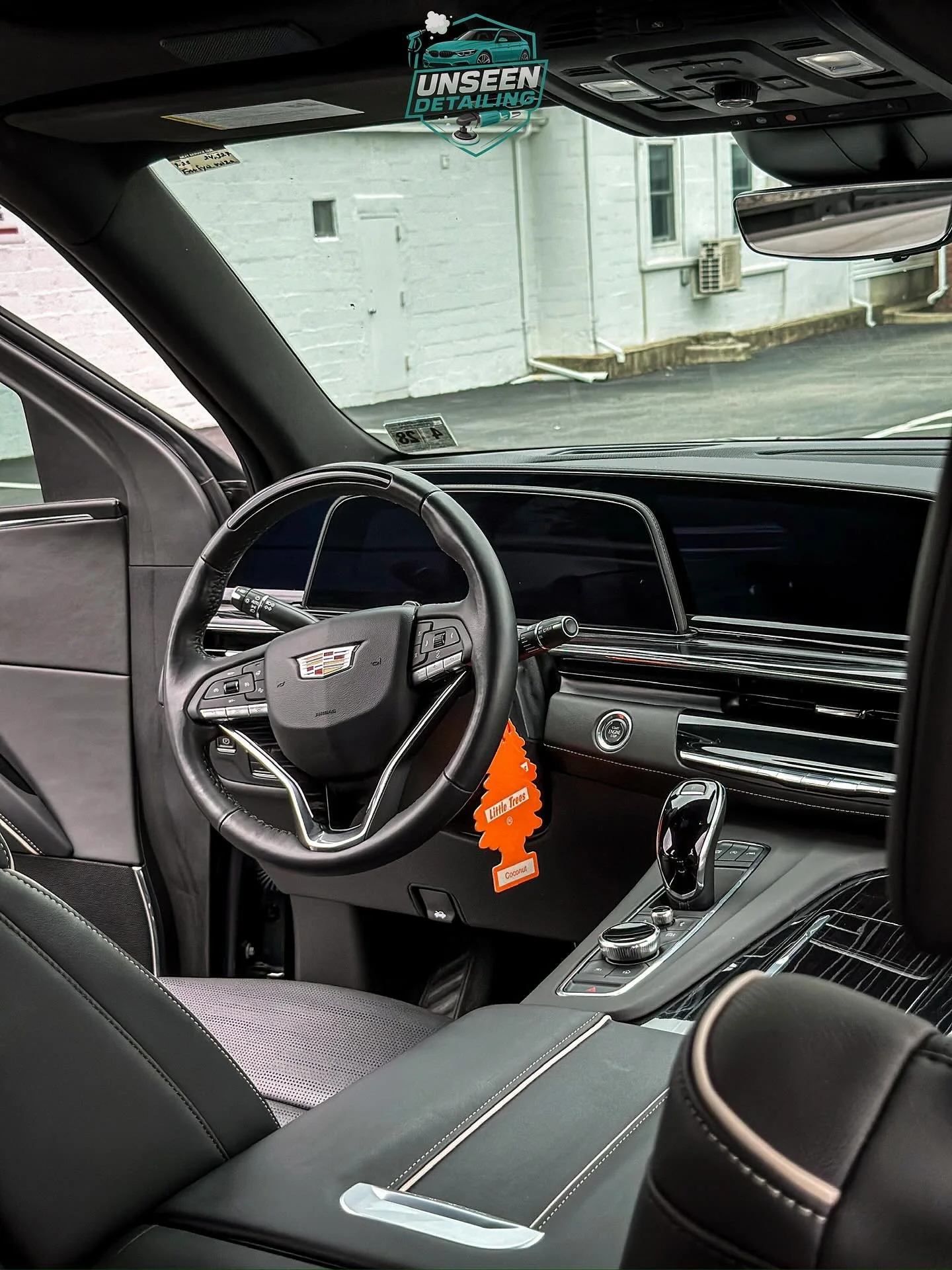 Interior of a Cadillac vehicle, showing the dashboard, steering wheel, gear shifter, and center console, with a building outside the windshield.