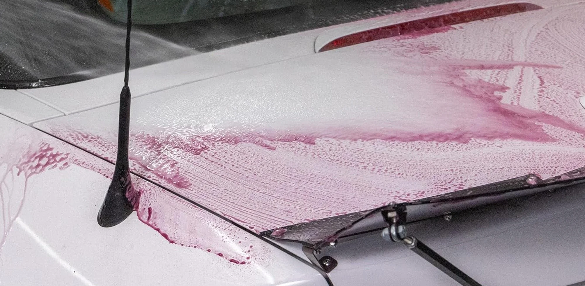 The hood of a white car covered in pink foam or soap suds, with a black antenna visible.