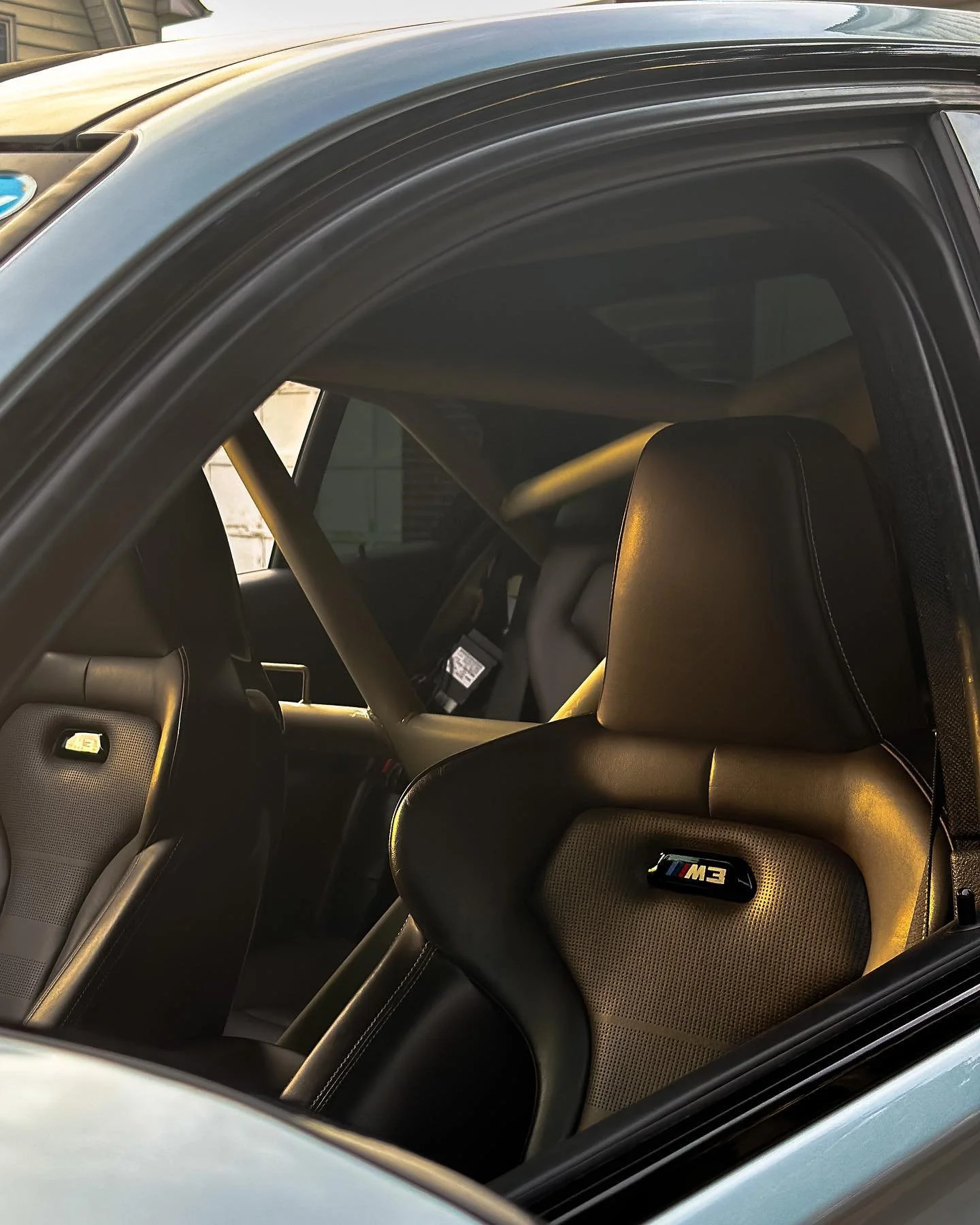 The interior of a BMW M3 sports car, showing black leather seats with a logo, and a roll cage installed in the backseat area.
