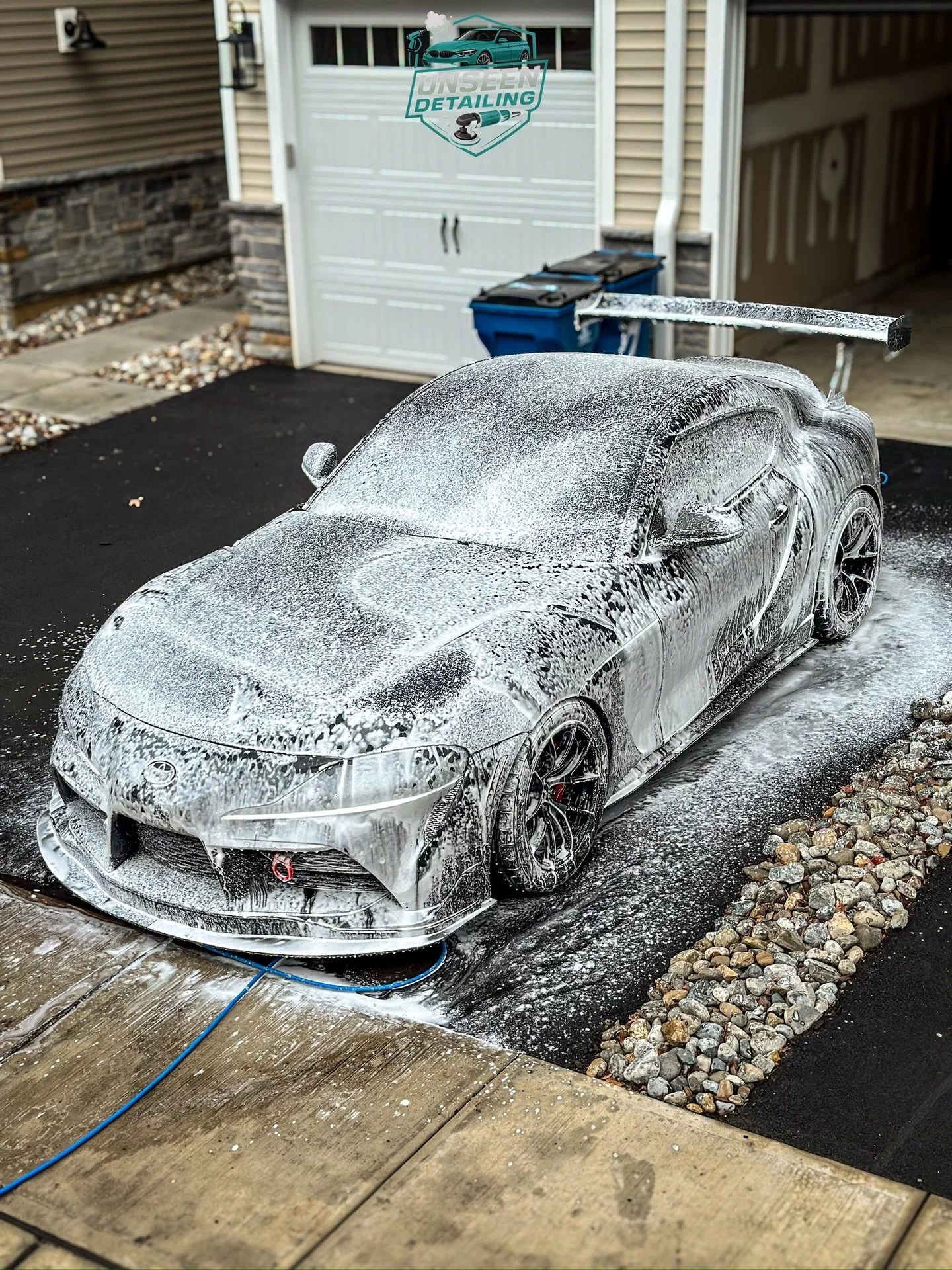 A detailed silver sports car is being washed with soap suds on the driveway of a house. The car has a large rear wing and black wheels, and is in the process of being cleaned.