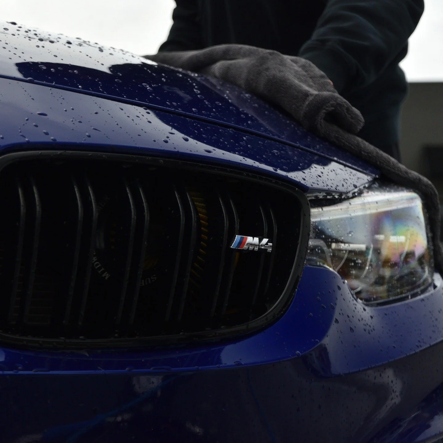 Close-up of the front of a blue BMW M4 car with water droplets on the hood and a person in black gloves polishing the car's surface.