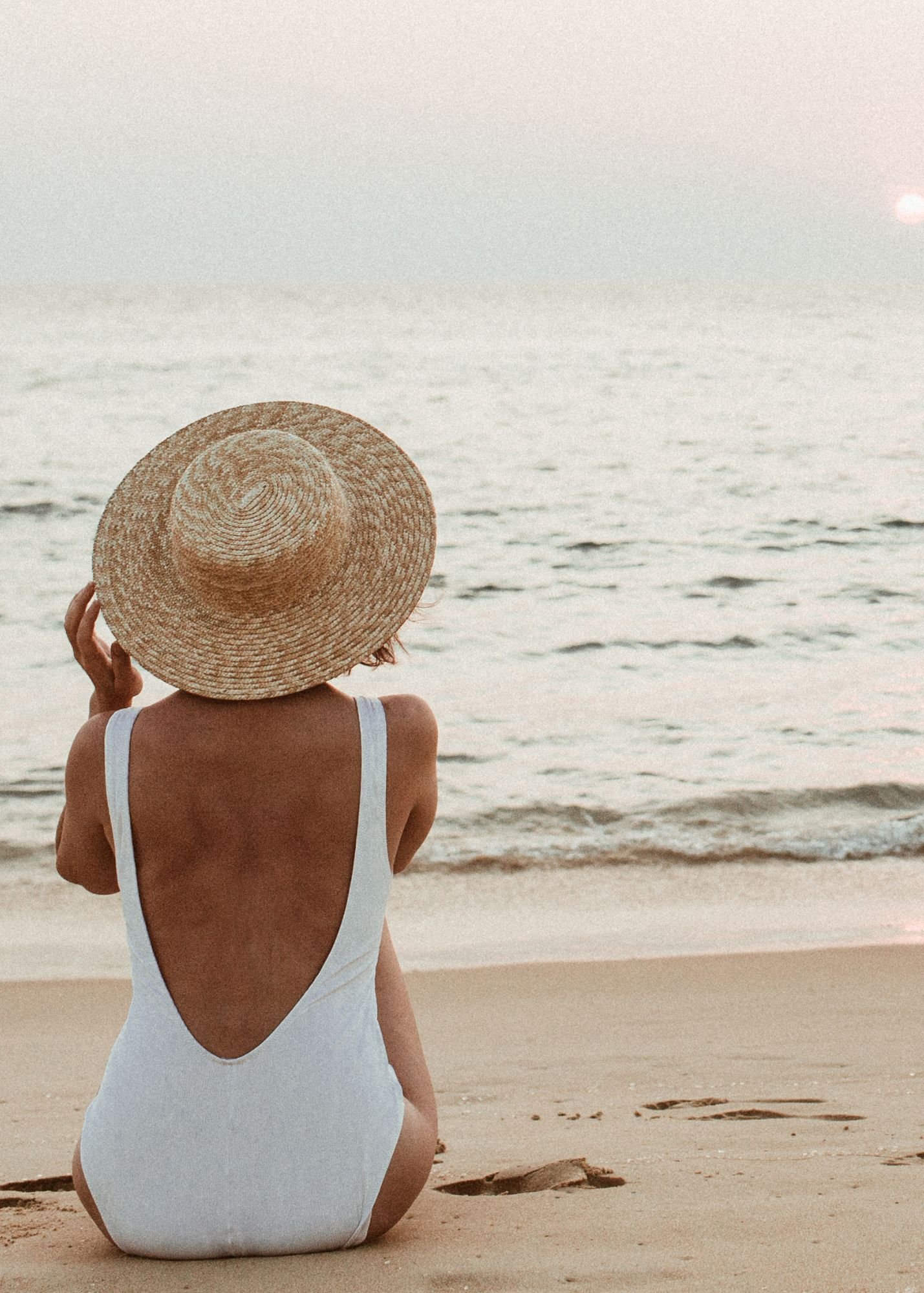 Woman sitting on the beach in a white swimsuit and a wide-brimmed straw hat, facing the ocean at sunset.