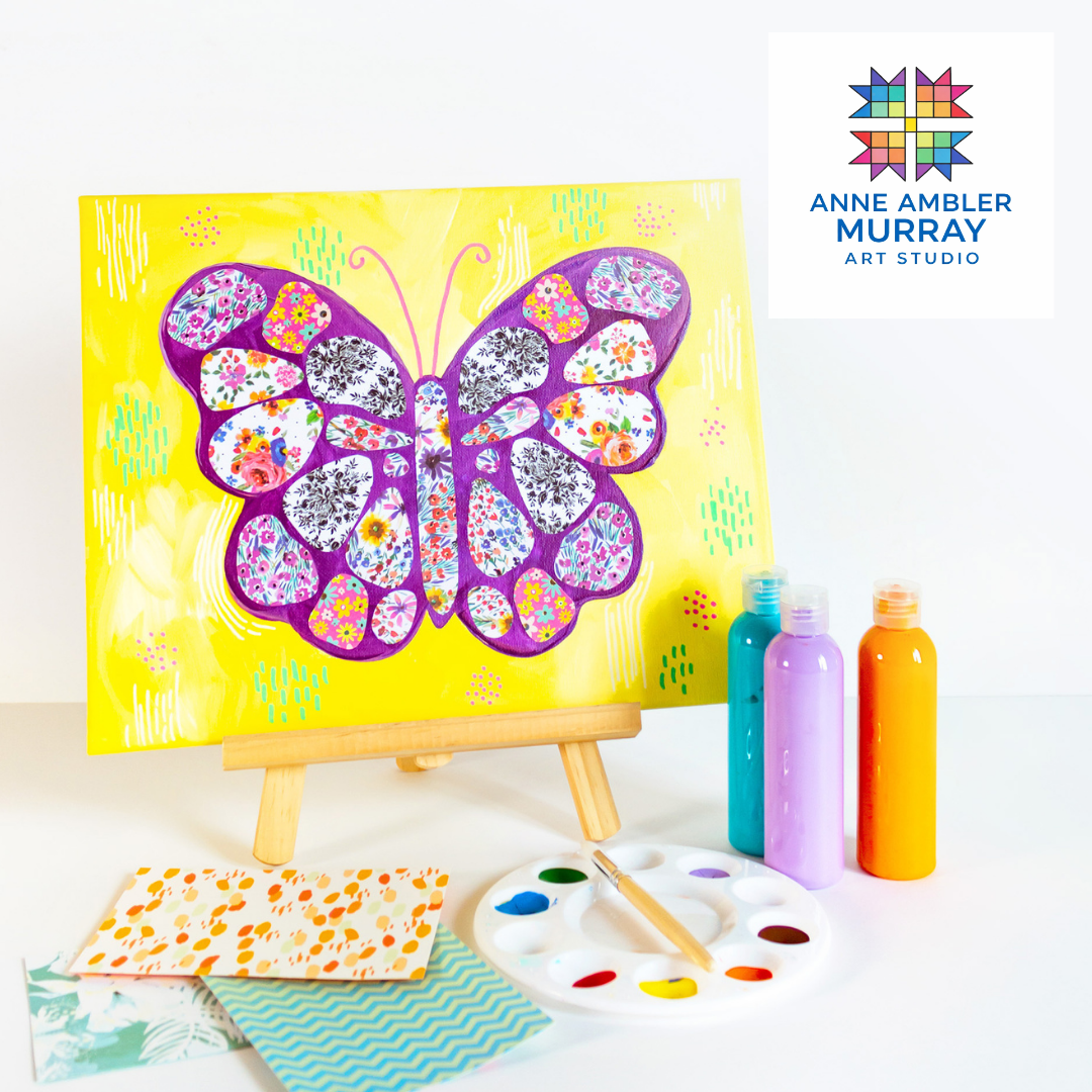 A colorful butterfly painting on a yellow background displayed on a small wooden easel, with bottles of paint, a paint palette, and patterned paper on a white surface.