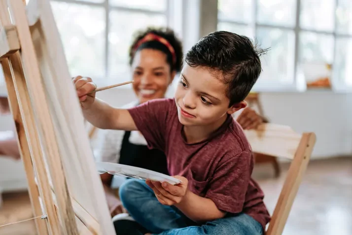 A young boy is painting on an easel in a bright, airy art studio, with a smiling woman in the background.