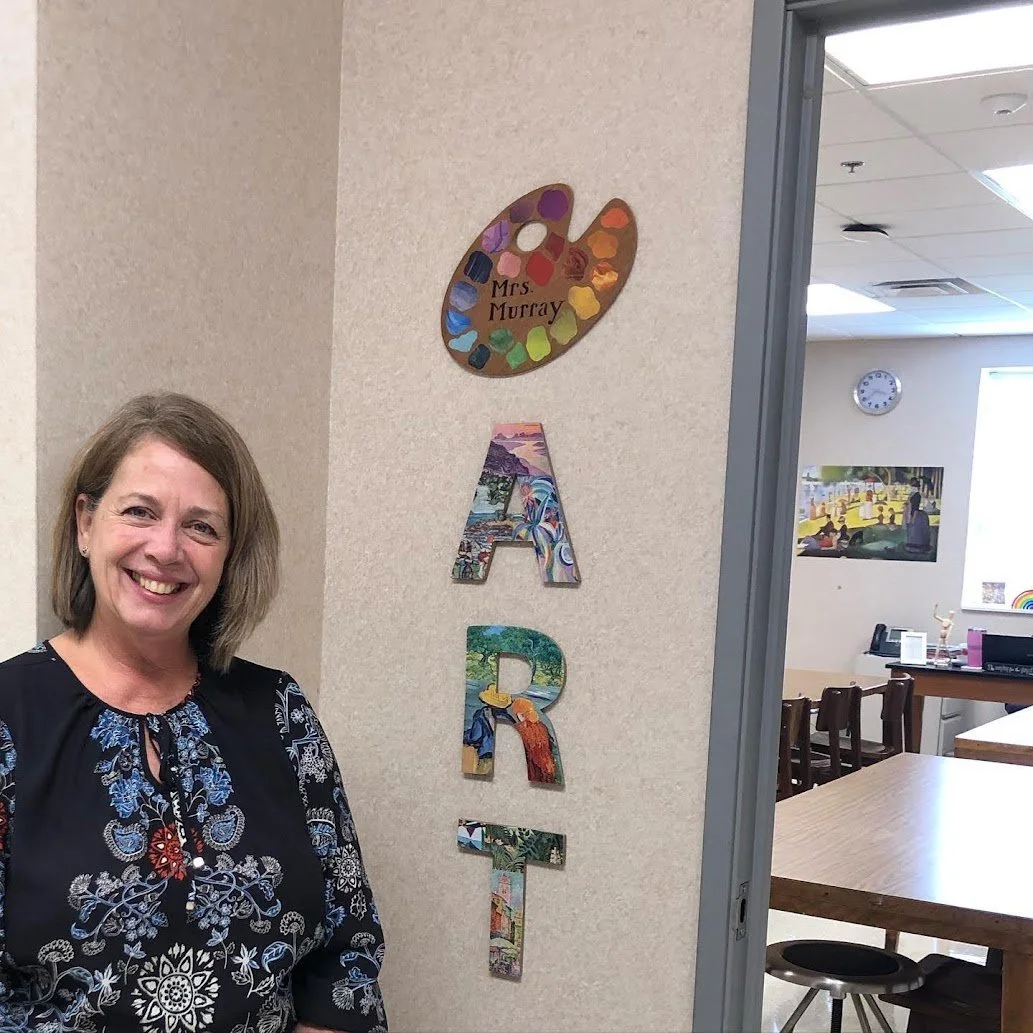 A smiling woman standing next to a wall with colorful letter cutouts spelling 'ART' and a painter's palette with the name 'Mrs. Murray' on it, in an art classroom or office.