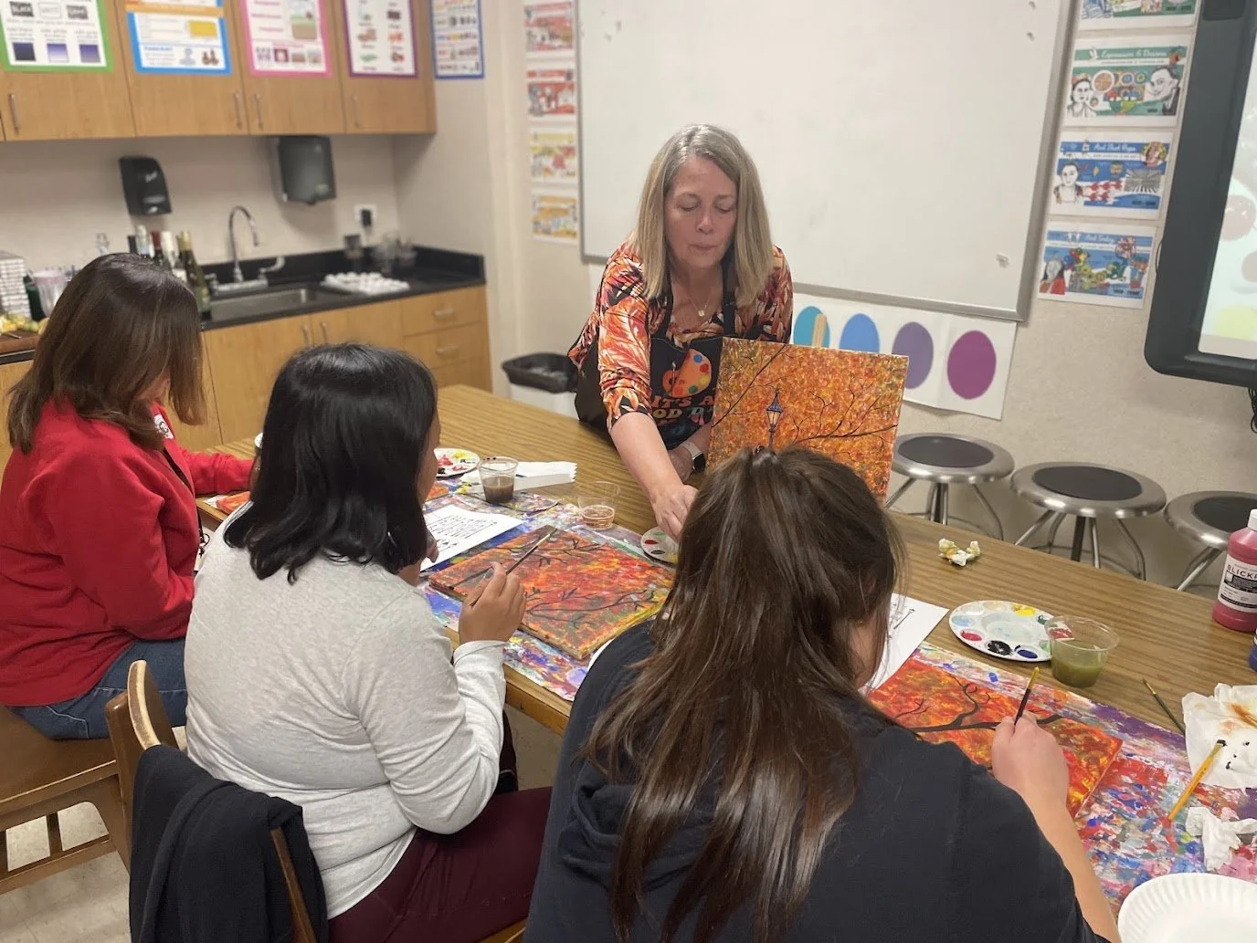 An art teacher demonstrating landscape painting with autumn trees to four students in a classroom.