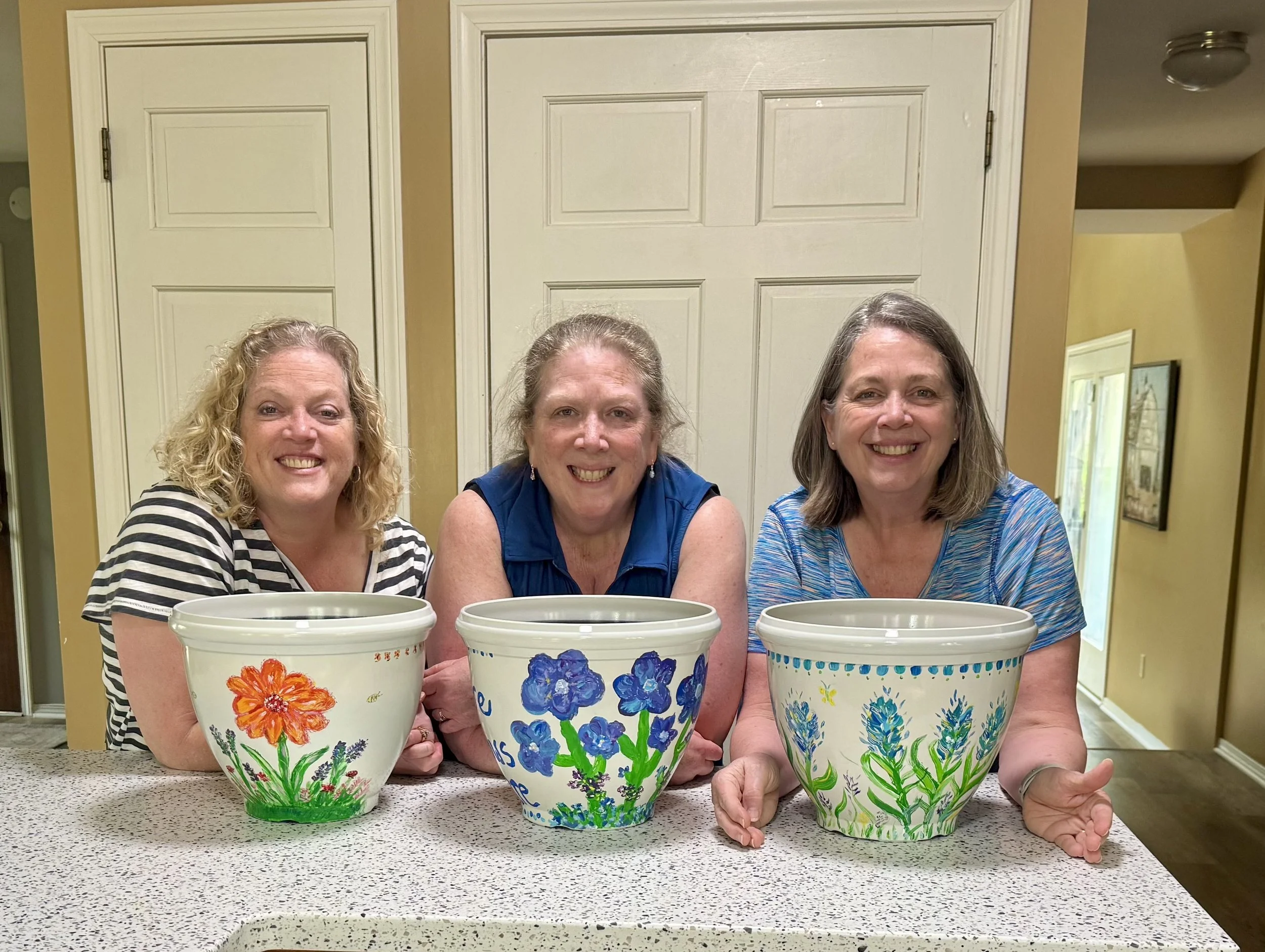 Three women smiling at the camera, each holding a large hand-painted flowerpot with colorful floral designs, sitting at a kitchen counter.