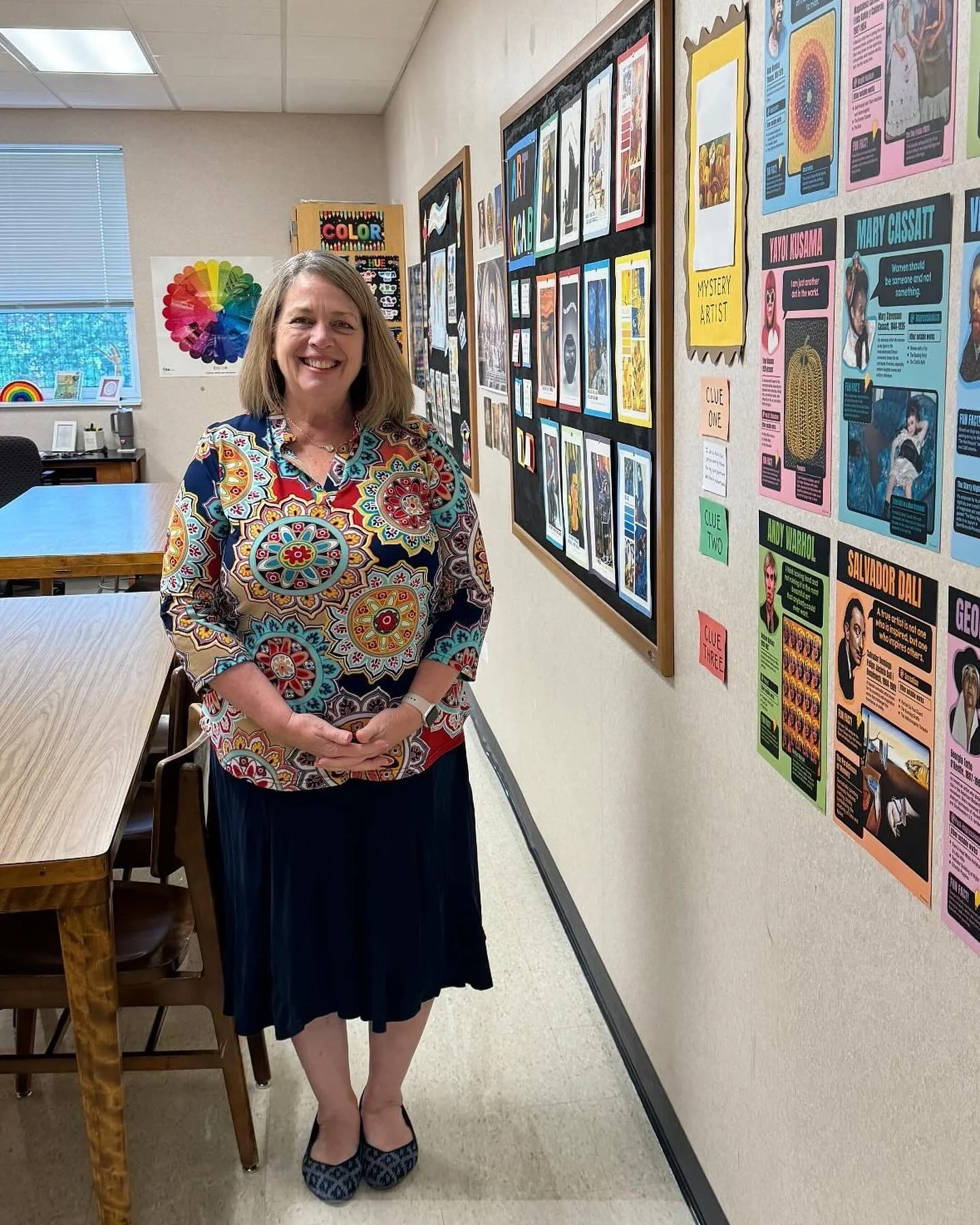 A woman standing in a classroom, smiling, wearing a colorful patterned blouse, a black skirt, and patterned shoes. She is in front of a bulletin board with educational posters and a wall with colorful displays.