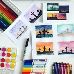 Colorful watercolor paintings of crosses at sunset, watercolor paints, brushes, and markers on a white desk.
