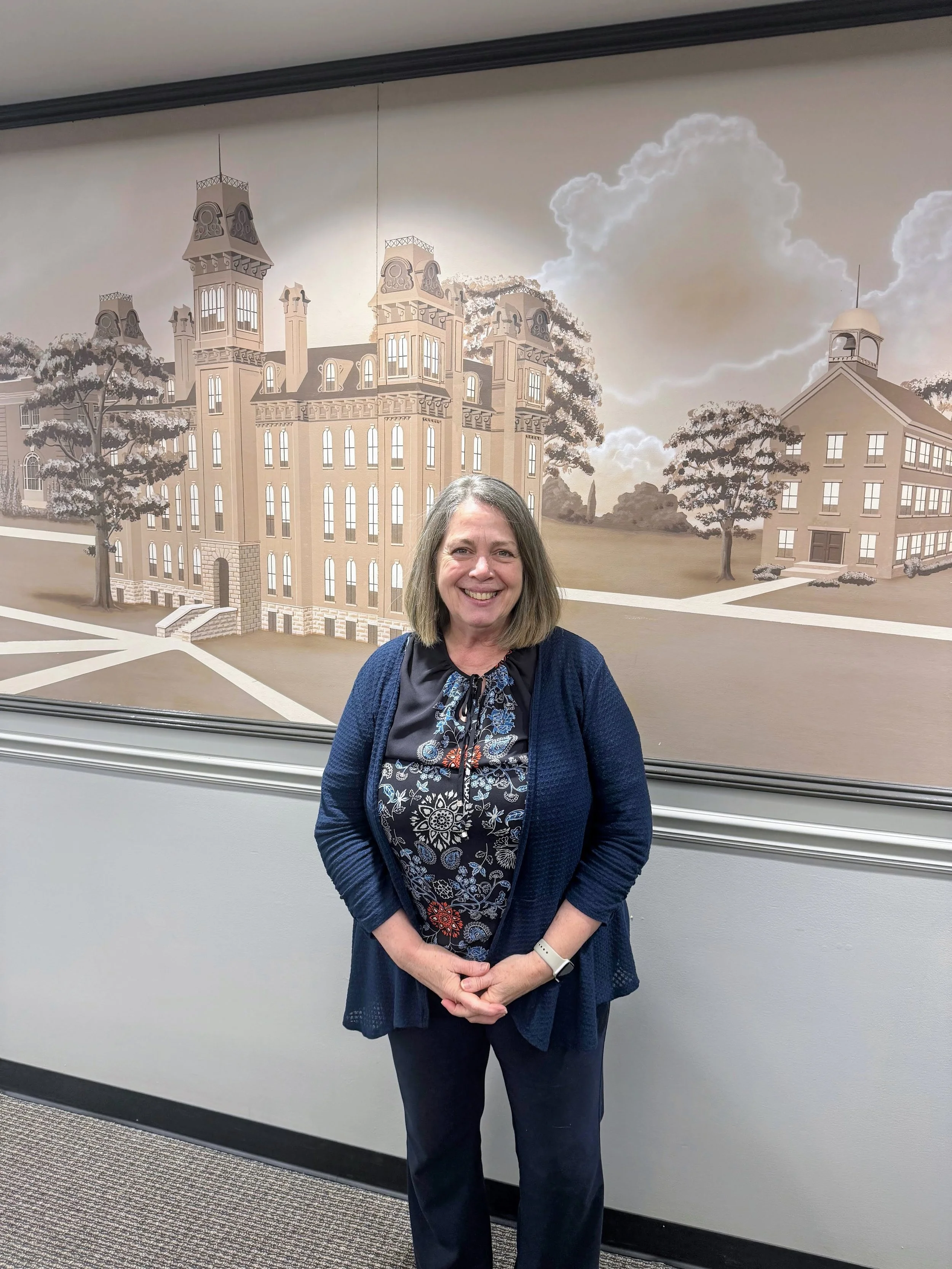 Smiling woman with shoulder-length hair standing in front of a mural depicting a historic building with towers and trees.