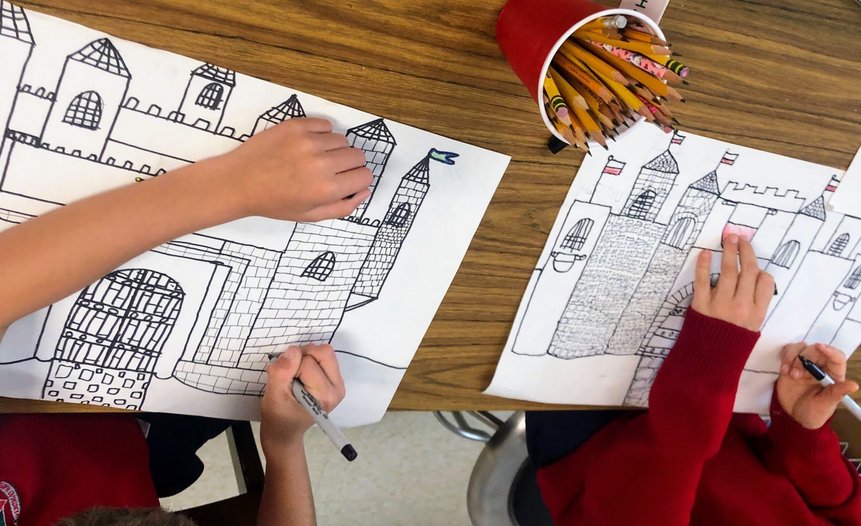 Two children are coloring castle drawings on white paper with black outlines, sitting at a wooden table
