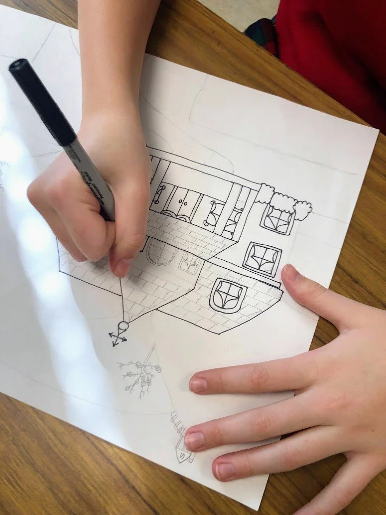 Child's hand drawing a house with a black marker on a sheet of paper, placed on a wooden table.